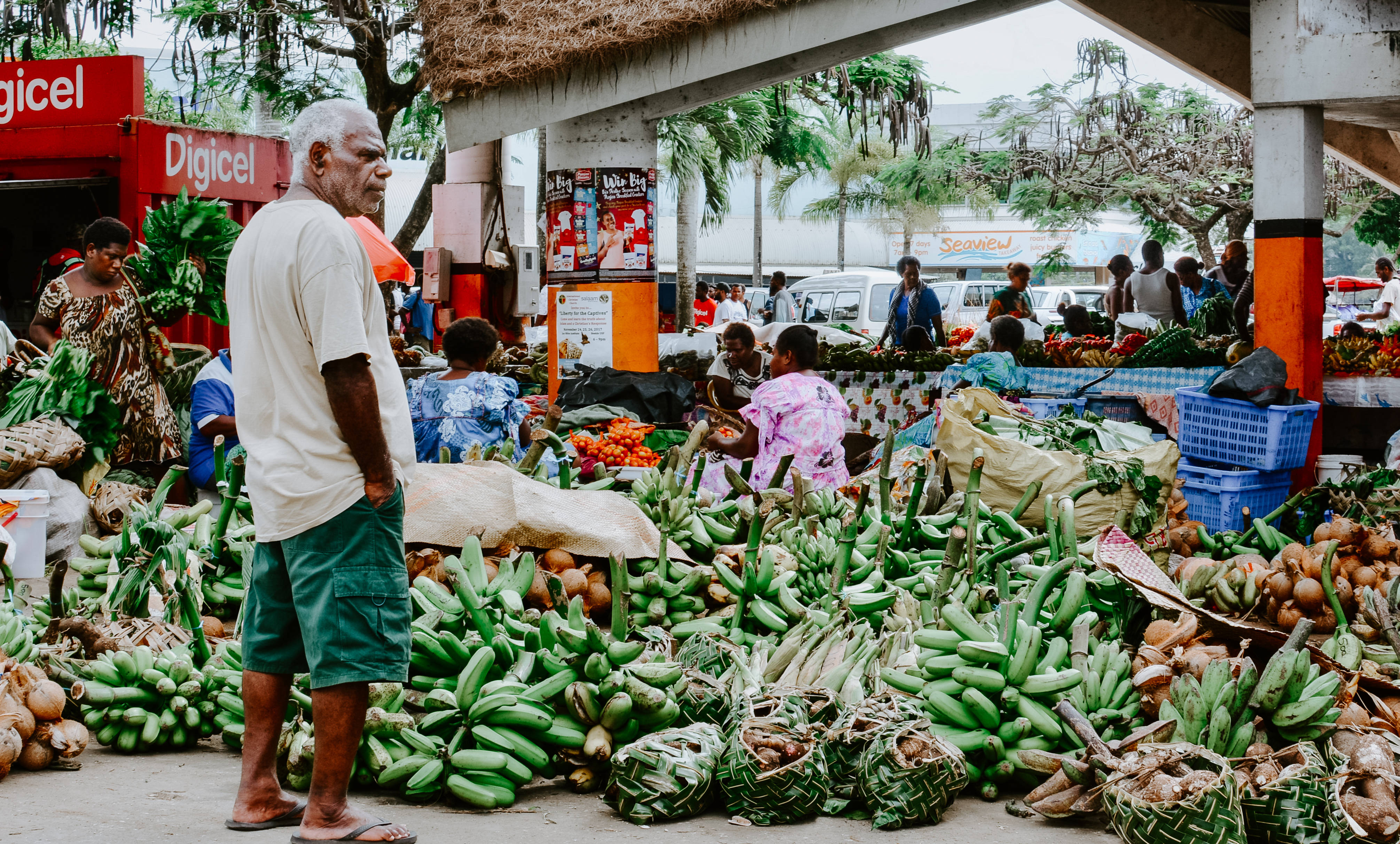 Vanuatu 桌布 壁紙 — 一個熱鬧的熱帶市場攤位，在茅草屋頂下堆滿了綠色香蕉和其他農產品，有購物者和攤販。