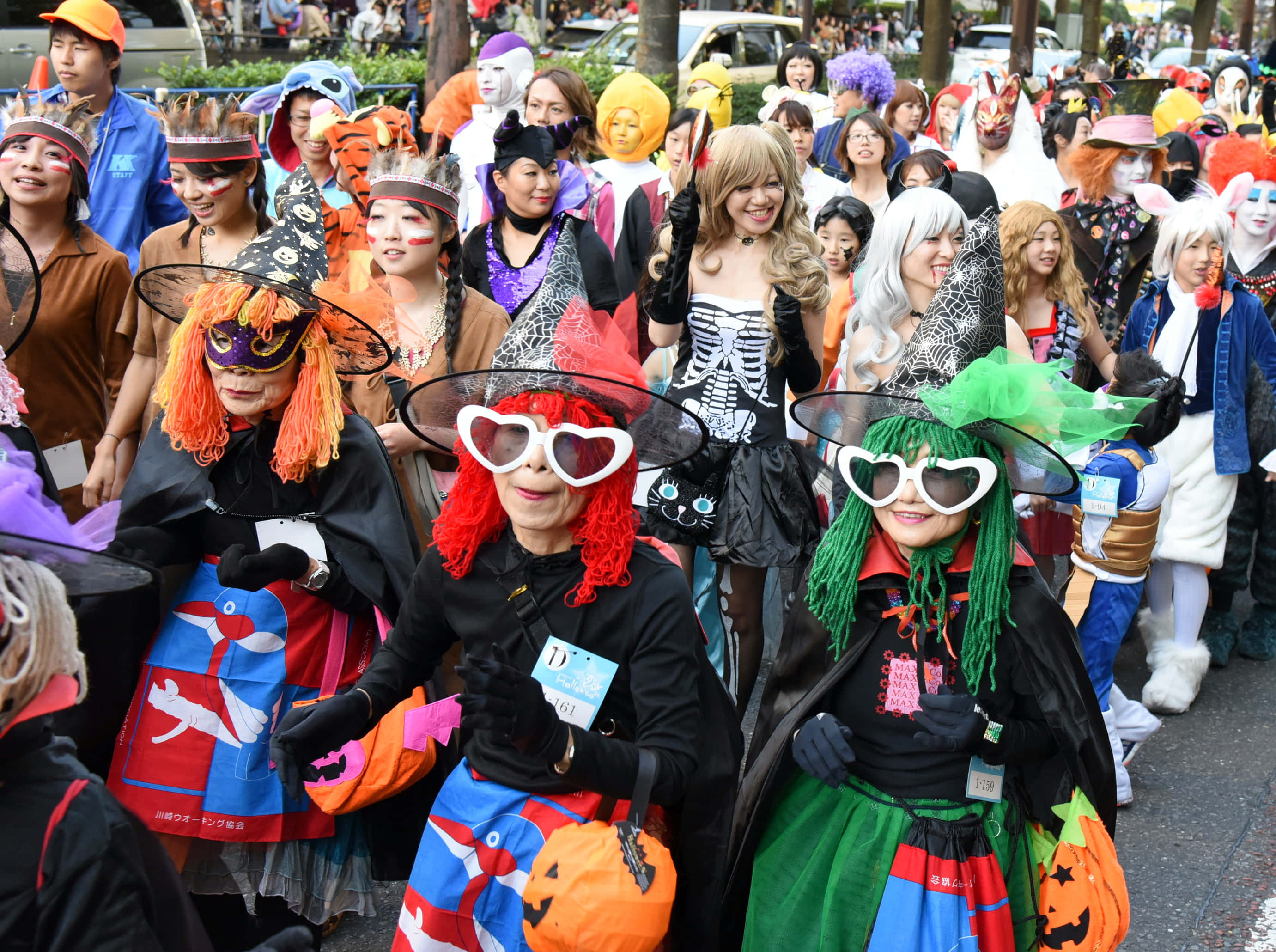 Halloween Parades 桌布 壁紙 — 一個熱鬧的萬聖節遊行，人們穿著色彩繽紛的服裝，在繁忙的街道上微笑並擺姿勢拍照。