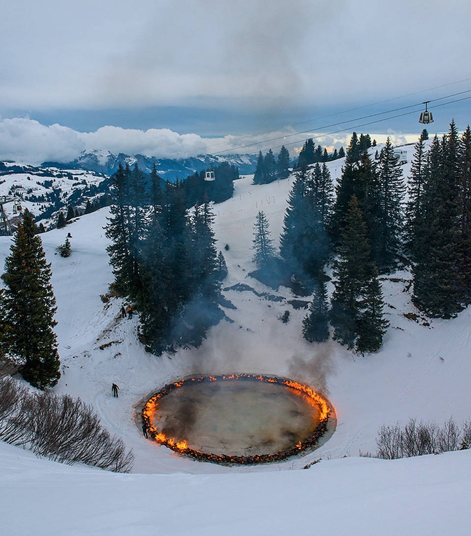 Alps 桌布 壁紙 — 一個熾熱的圓形水池在白雪覆蓋的山谷中明亮地燃燒，周圍環繞著常青樹和滑雪纜車，天空多雲。
