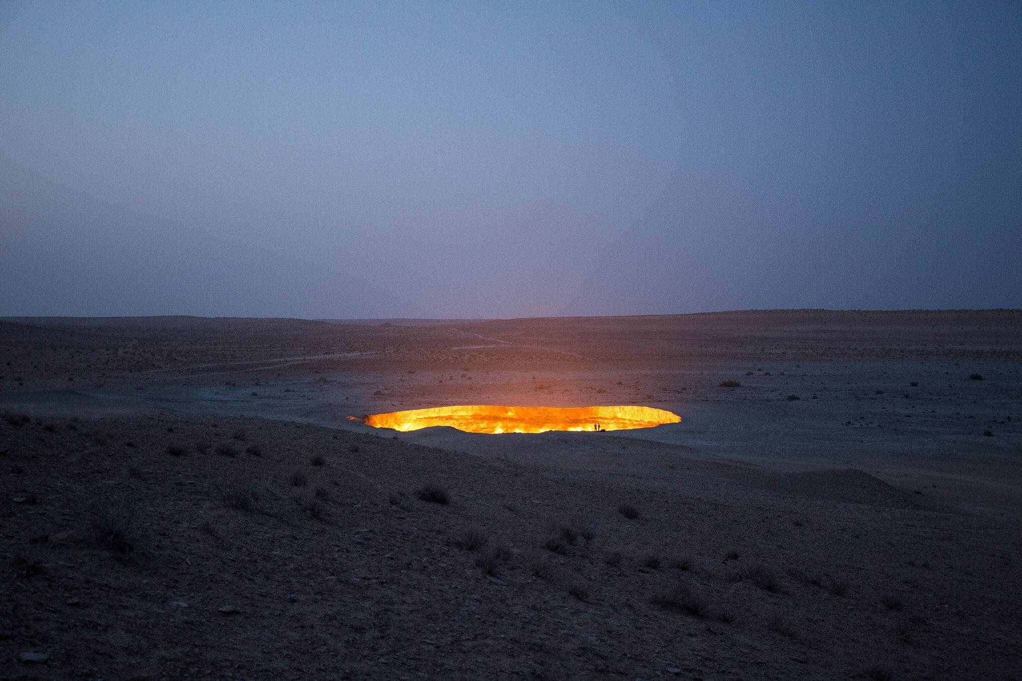 Turkmenistan 桌布 壁紙 — 一個熾熱的橙色氣體火山口在暮色天空下的廣闊黑暗沙漠中明亮地燃燒。