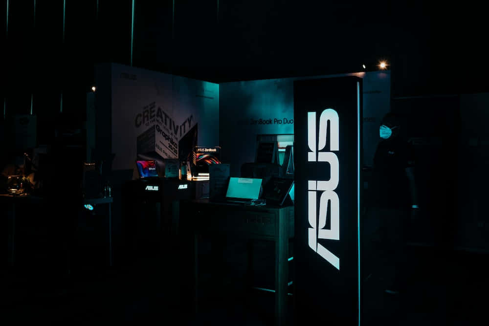 ASUS 桌布 壁紙 — 一個燈光昏暗的ASUS科技展位，帶有發光的藍色標誌和展示中的筆記型電腦，位於黑暗的展覽廳內。