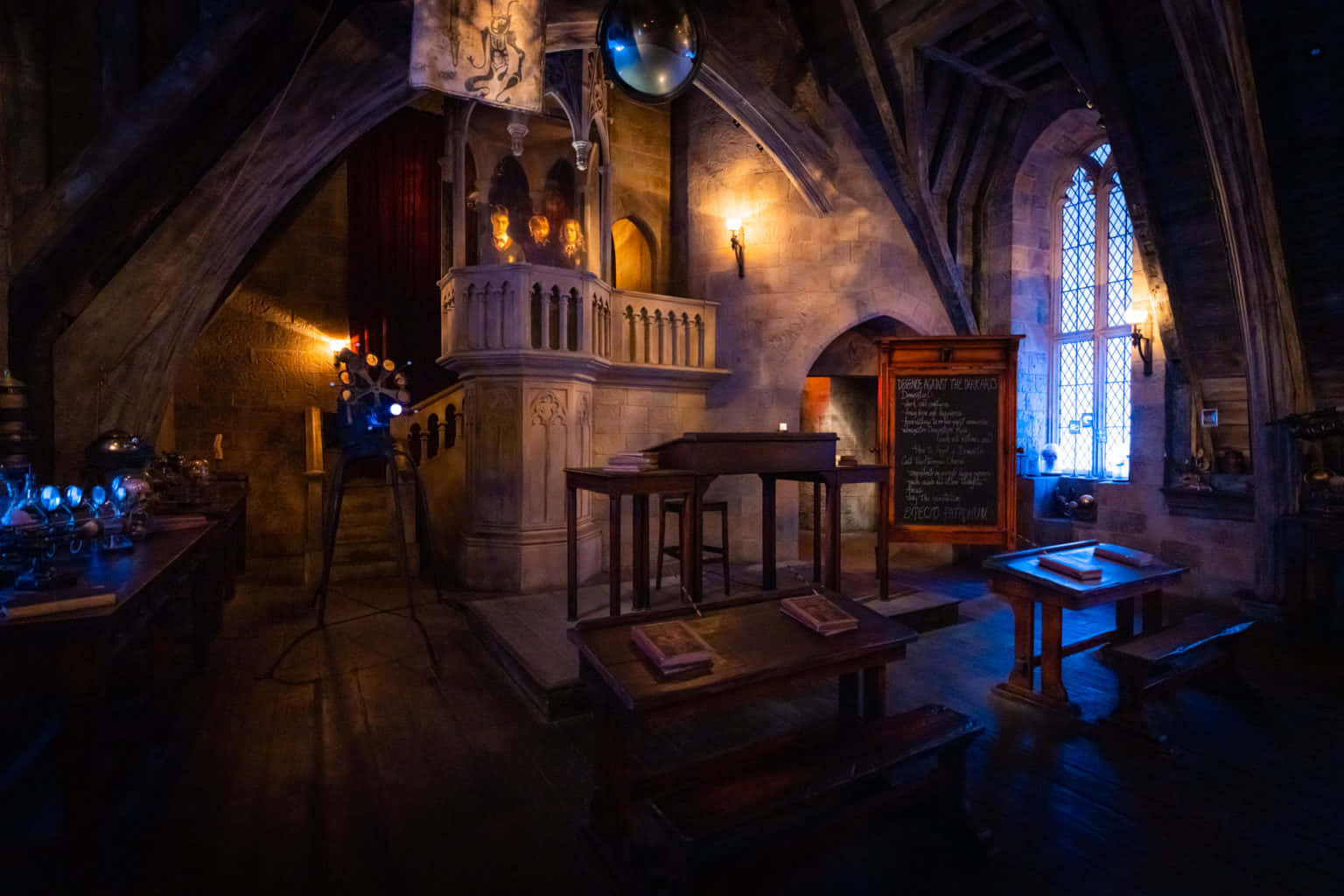 The Hogwarts Dark Arts Classroom 桌布 壁紙 — 一個燈光昏暗的中世紀風格煉金術實驗室，配有木桌、發藍光的藥水、石牆和拱形窗戶，投射出溫暖的光線。