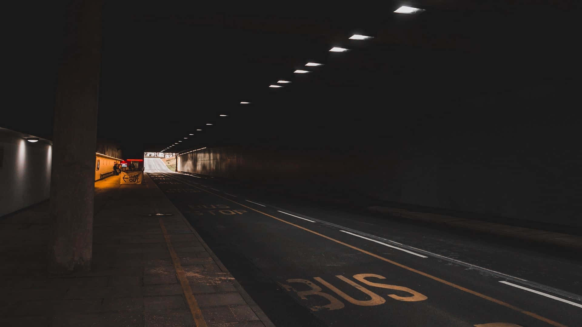 Dark Underground 桌布 壁紙 — 一個燈光昏暗的城市隧道，頭頂燈光在道路上標記的公車站上投下長長的陰影。