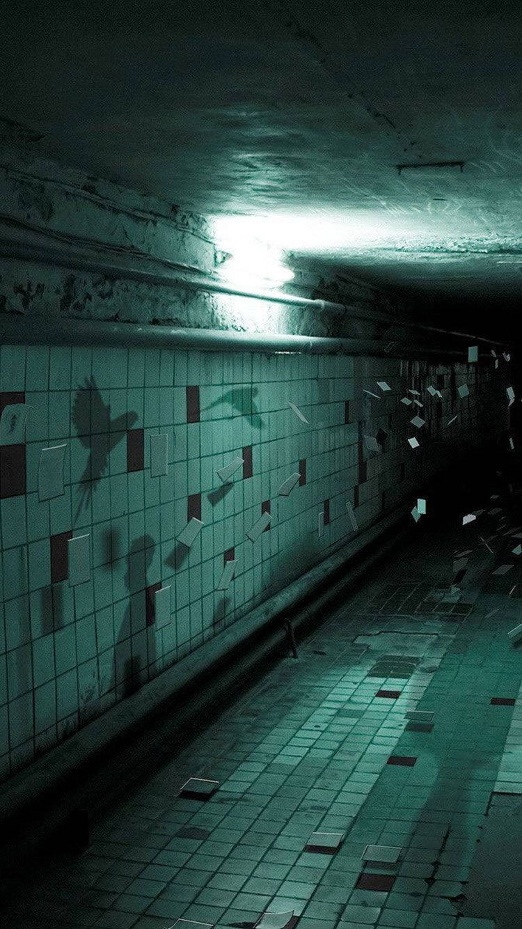 Scary Halloween iPhone 桌布 壁紙 — 一個燈光昏暗的廢棄地鐵隧道，裂開的瓷磚牆壁和地板，由單一頭頂燈泡照亮，投射出詭異的陰影。