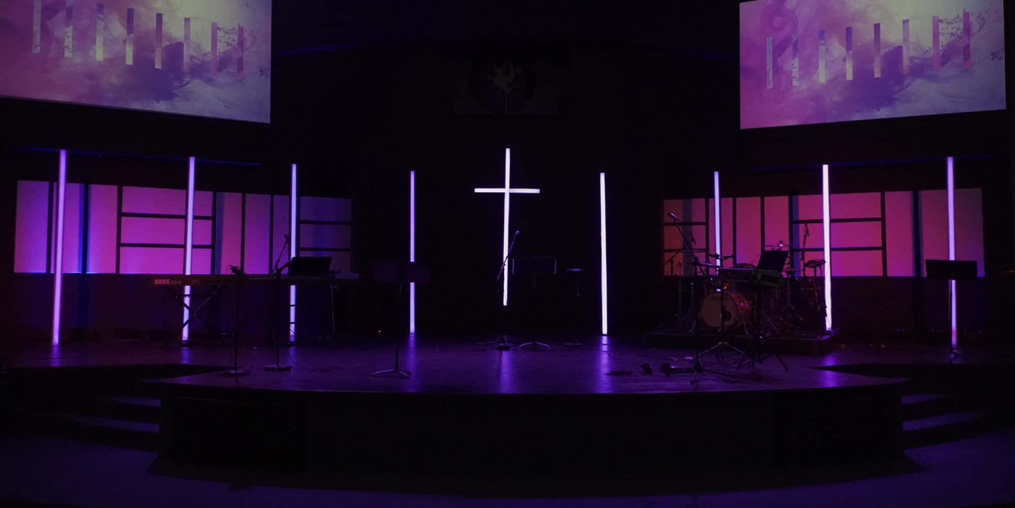 Church Stage 桌布 壁紙 — 一個燈光昏暗的教堂舞台，沐浴在紫色燈光中，中央有一個發光的十字架，樂器準備好進行敬拜。
