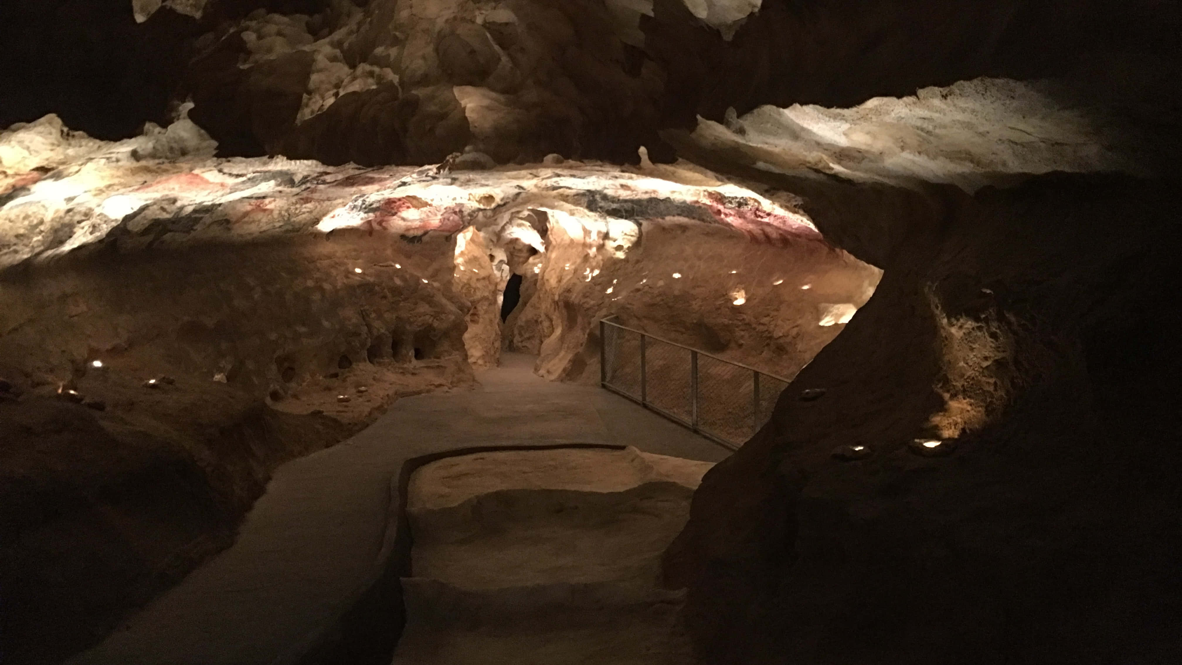 Lascaux Caves 桌布 壁紙 — 一個燈光昏暗的洞穴，沿著蜿蜒的步道，岩壁上古老的紅色赭石畫作被聚光燈照亮。