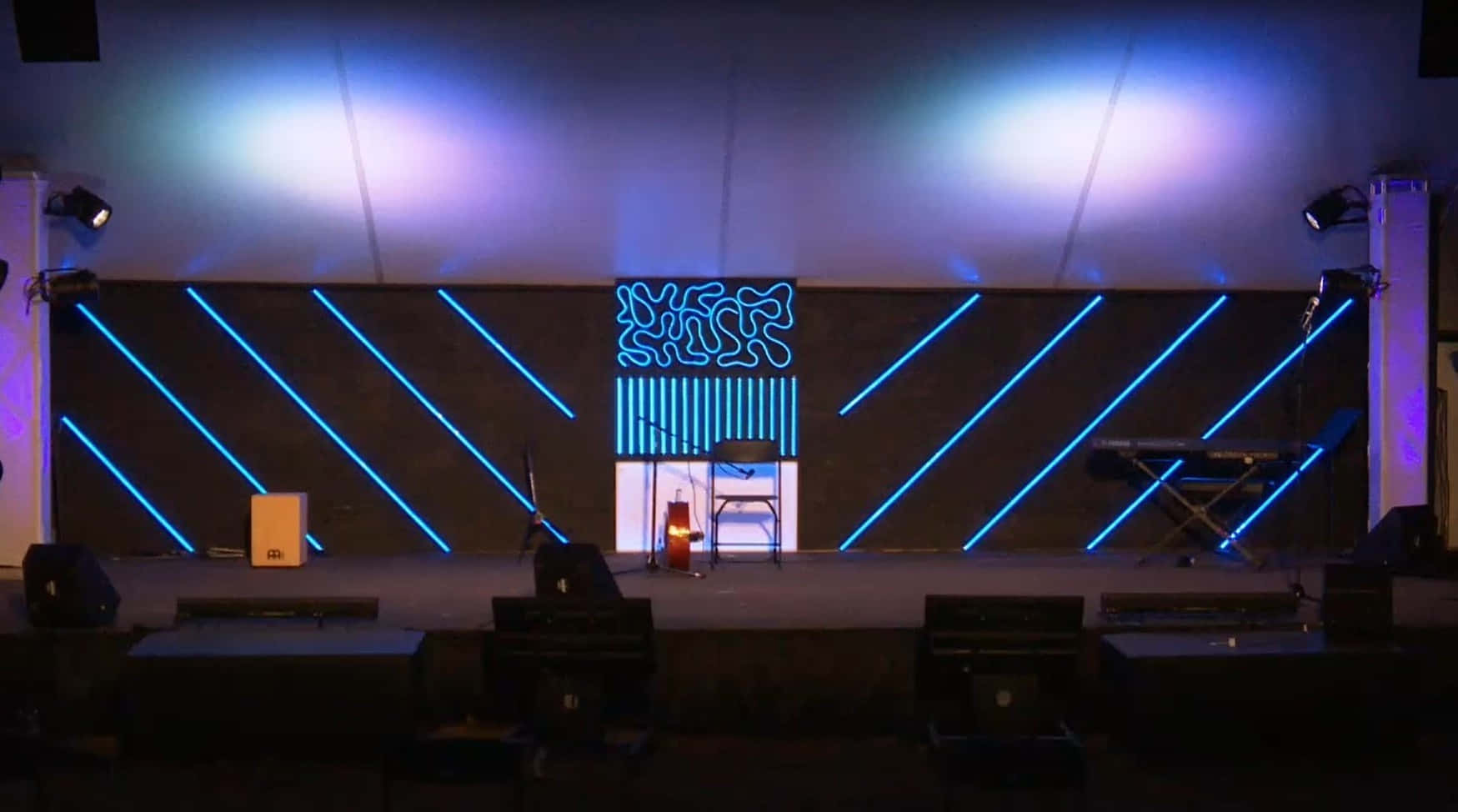 Church Stage 桌布 壁紙 — 一個燈光昏暗的舞台，帶有藍色霓虹燈和背景上發光的抽象圖案。