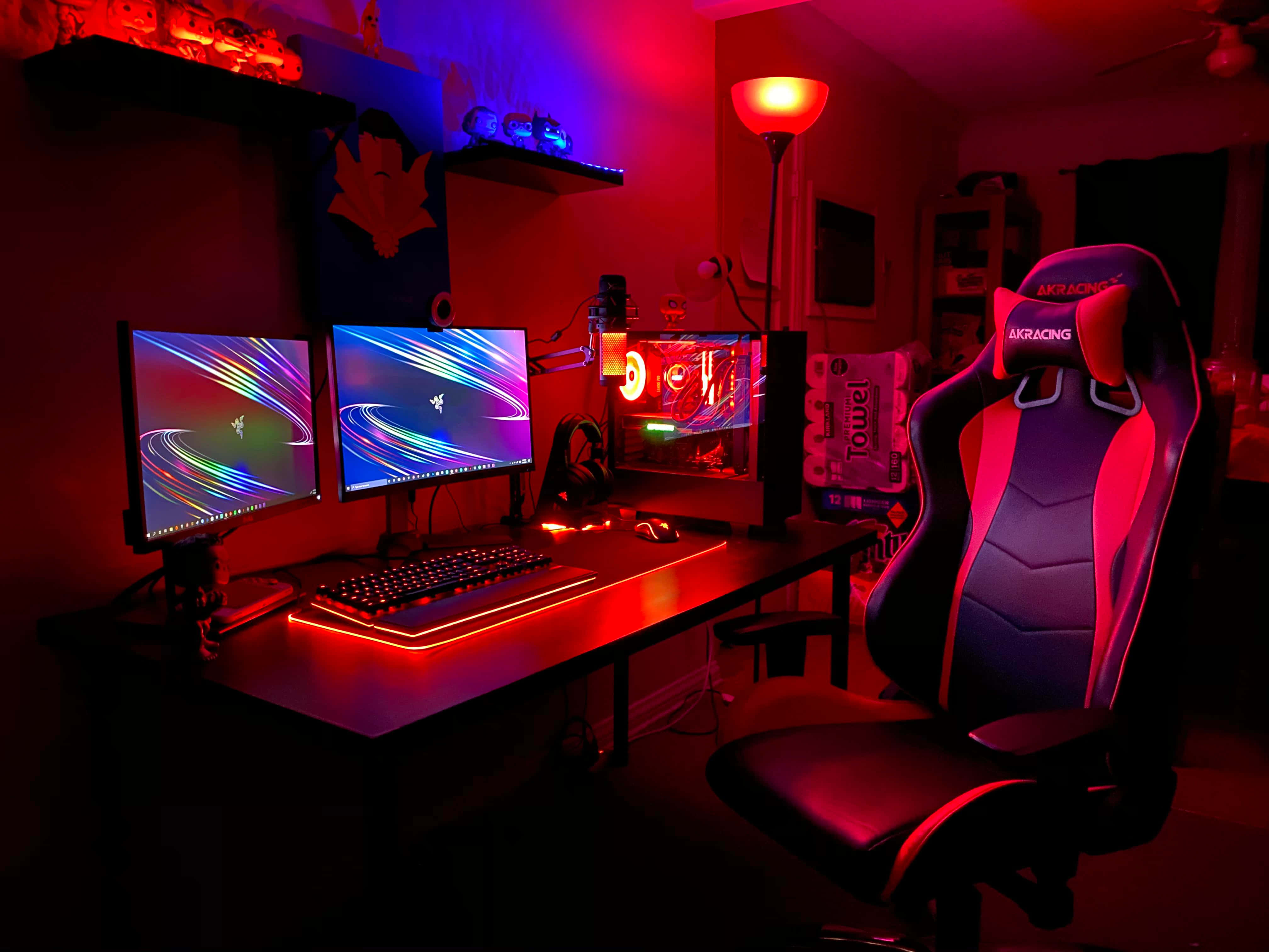 Gaming Pc Setup 桌布 壁紙 — 一個燈光昏暗的遊戲設置，沐浴在紅色和藍色LED光芒中，配備多個顯示器和一張賽車椅。