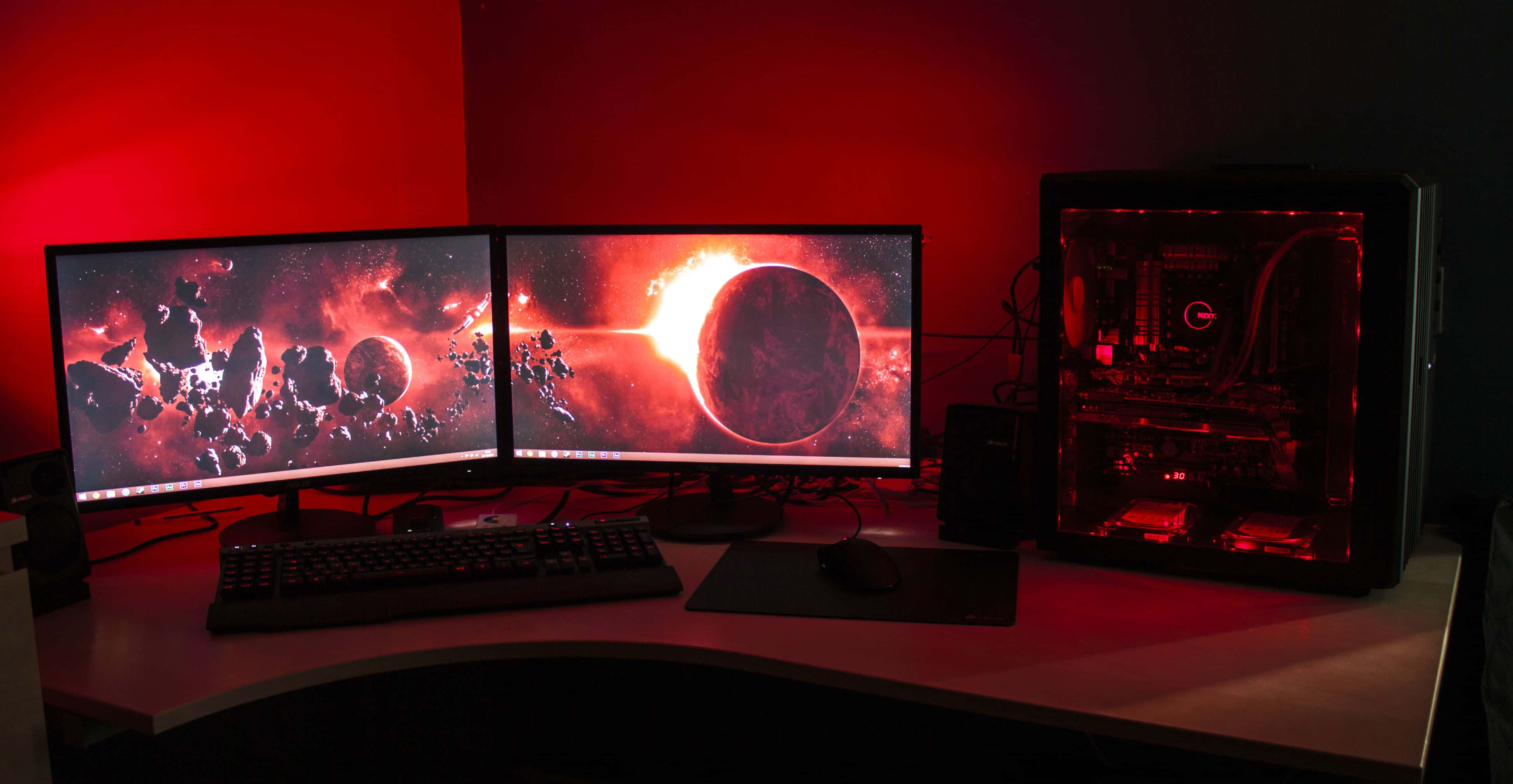 Gaming Setup 桌布 壁紙 — 一個燈光昏暗的遊戲設置，配備雙顯示器顯示熾熱的宇宙場景，發光的紅色PC機殼在桌子上投射環境光。