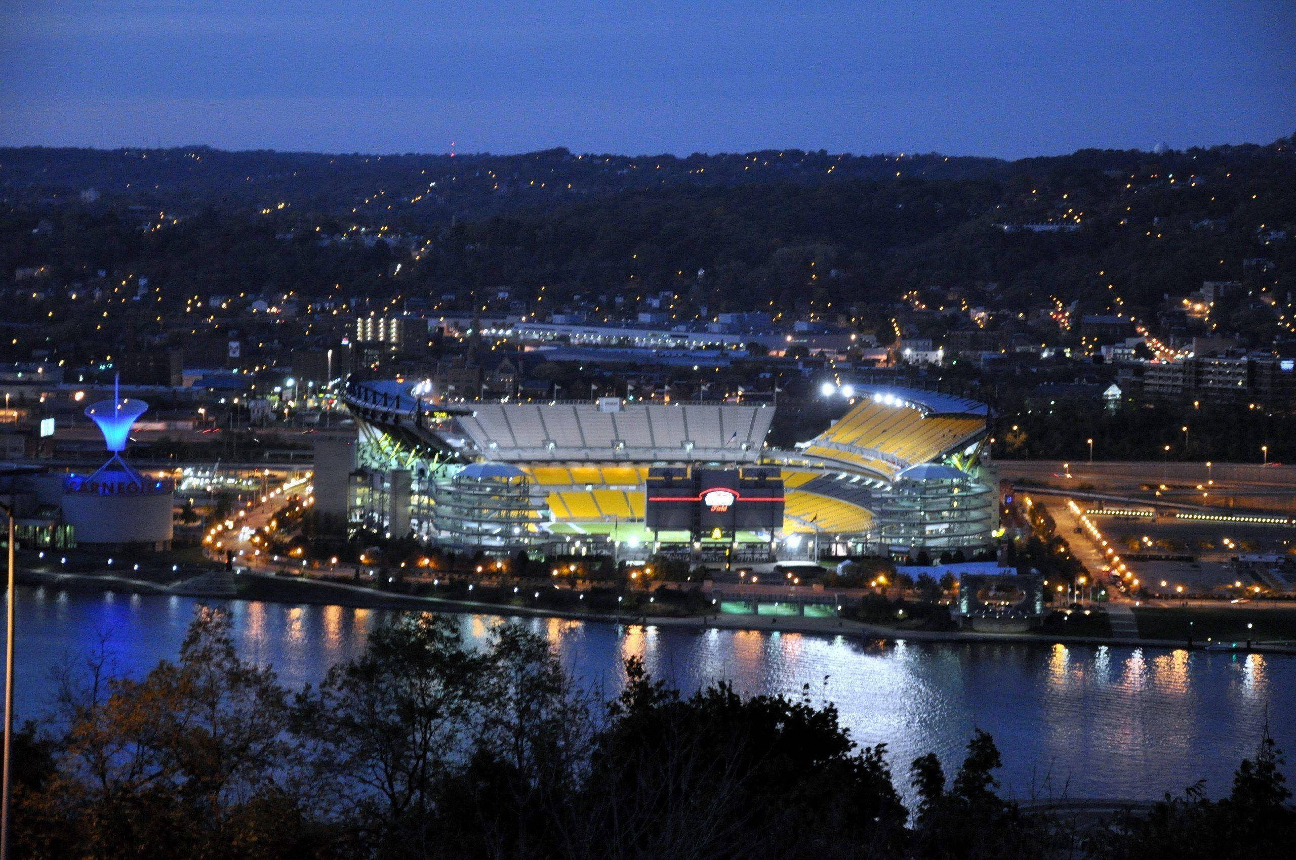 夜空 桌布 壁紙 — 一個燈火通明、設有黃色座位的體育場在黃昏時分在河邊閃耀，河水倒映著城市燈光與山丘。
