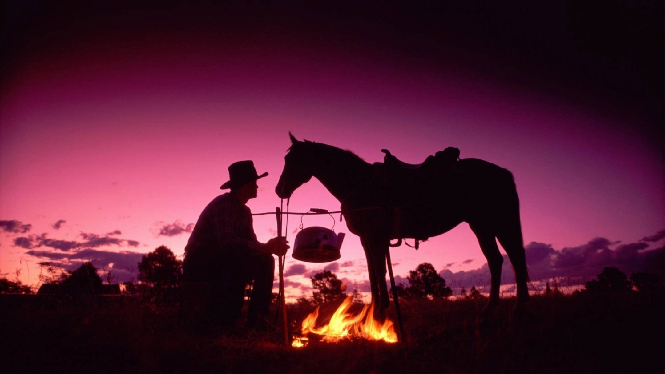 Cowboy 桌布 壁紙 — 一個牛仔在鮮豔的紫色日落襯托下形成剪影，正在營火旁為他備好鞍的馬煮水。