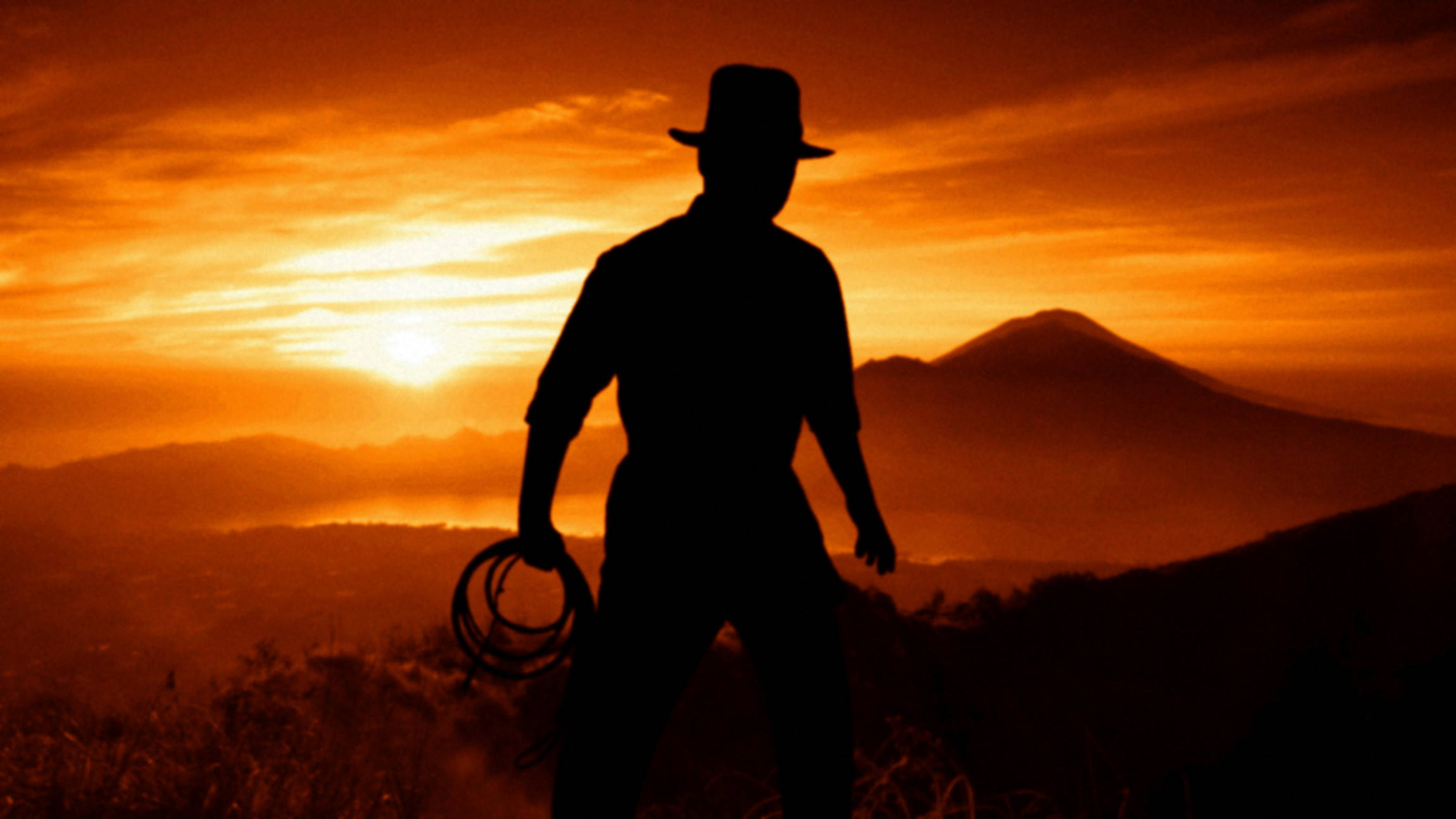 Indiana Jones 桌布 壁紙 — 一個牛仔手持套索的剪影，站在遠山之上火紅的橙色日落前。
