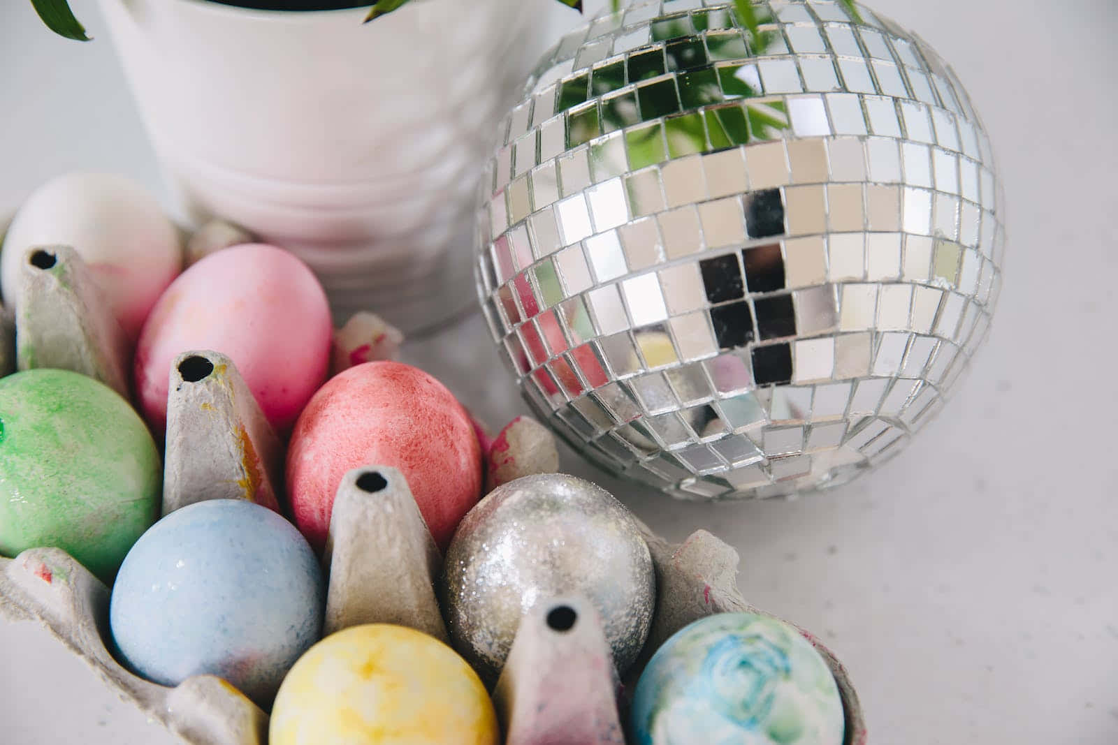 Pastel Easter 桌布 壁紙 — 一個特寫，色彩繽紛的手繪雞蛋放在紙板托盤中，旁邊是一個閃亮的鏡面迪斯科球和一個白色盆栽植物。