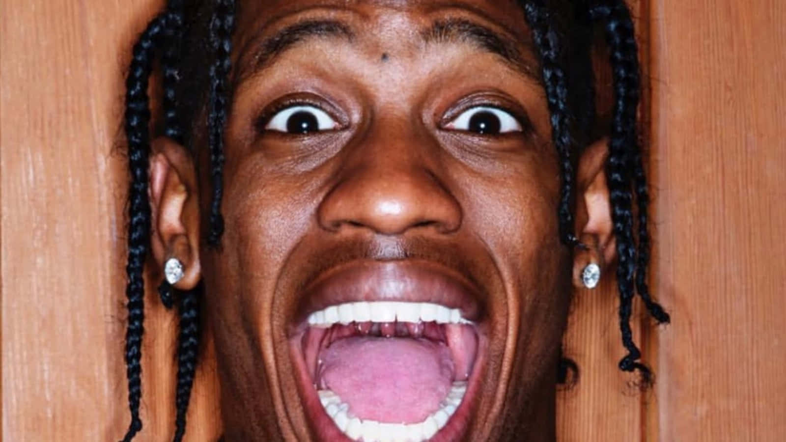 Travis Scott Funny 桌布 壁紙 — 一個特寫照片，一個男人編著辮子，戴著鑽石耳環，對著木質背景睜大眼睛尖叫。