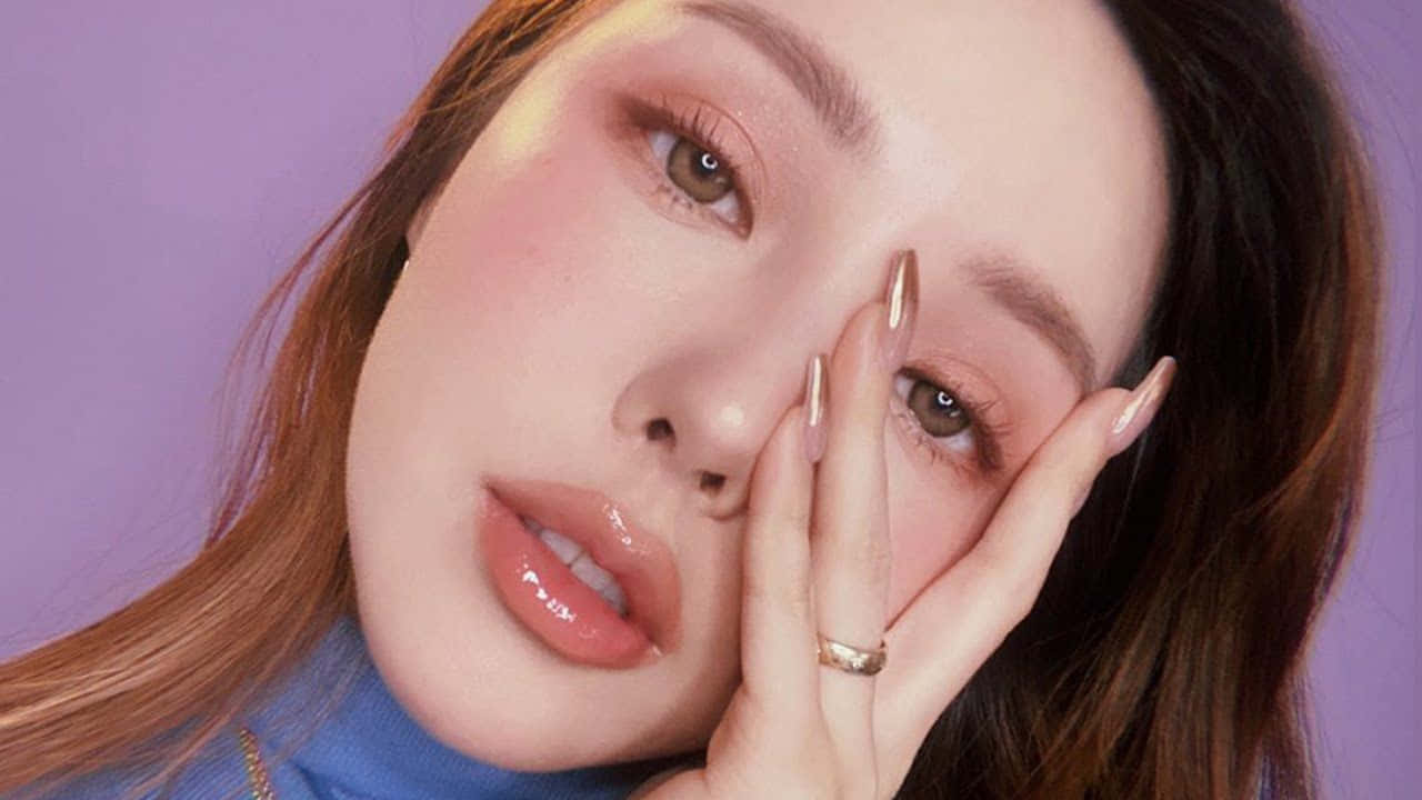 Spring Makeup 桌布 壁紙 — 一個特寫肖像，一位化著淡妝、留著長指甲的女性，在紫色背景前輕輕觸摸自己的臉。