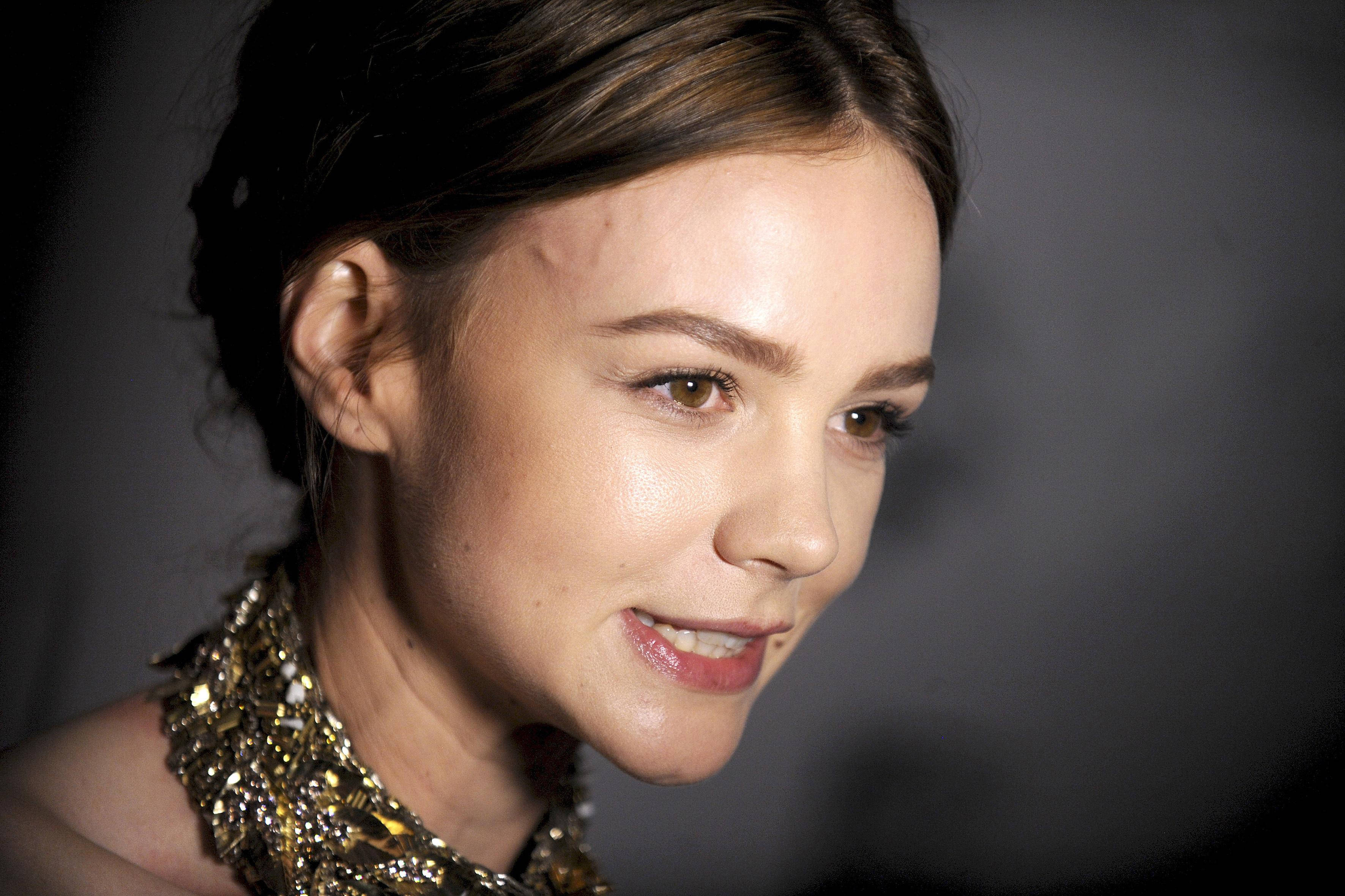 Carey Mulligan 桌布 壁紙 — 一個特寫肖像，一位擁有棕色頭髮和金色珠鏈的女性，背景為深灰色。