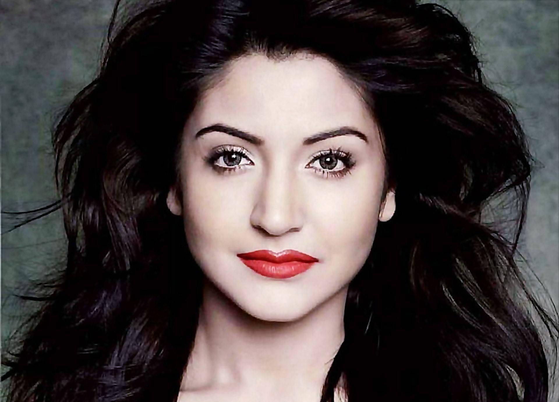 Anushka Sharma 桌布 壁紙 — 一個特寫肖像，一位擁有深色波浪頭髮和大膽紅唇的女性，背景是柔和的灰色。