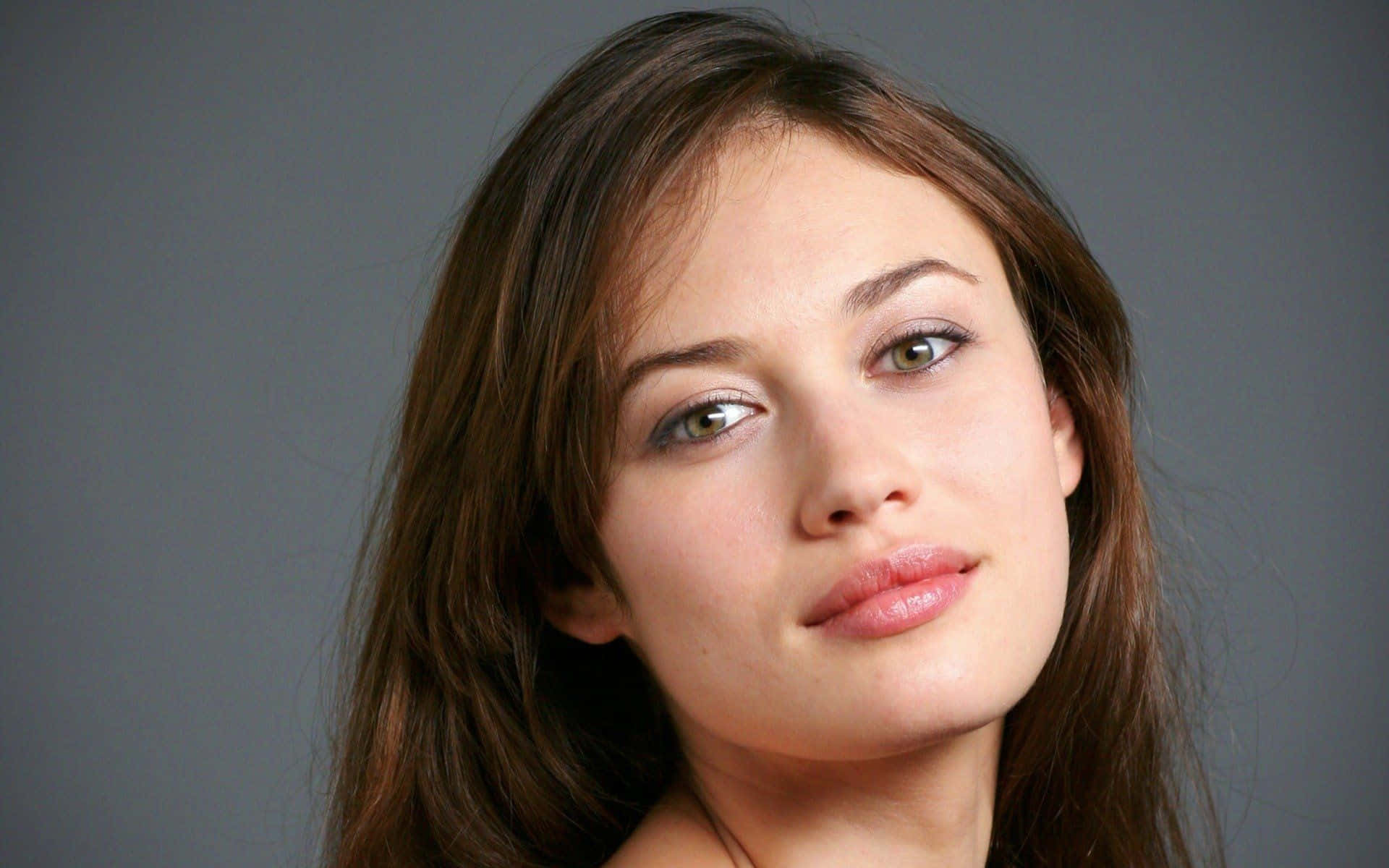 Olga Kurylenko 桌布 壁紙 — 一個特寫肖像，一位棕色頭髮、綠色眼睛的女性在灰色背景前直視鏡頭。