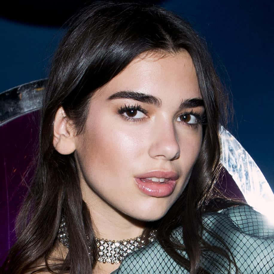 Dua Lipa 桌布 壁紙 — 一個特寫肖像，一位深色頭髮、妝容大膽的女性，戴著鑲有寶石的頸鏈和網狀上衣，背景是充滿活力的紫色和藍色。