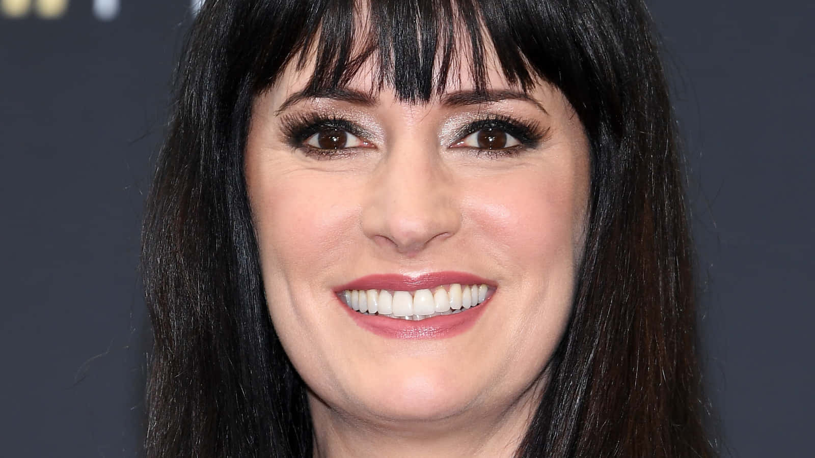 paget brewster 桌布 壁紙 — 一個特寫肖像，一位深色頭髮和瀏海的女性在深灰色背景前溫暖地微笑。