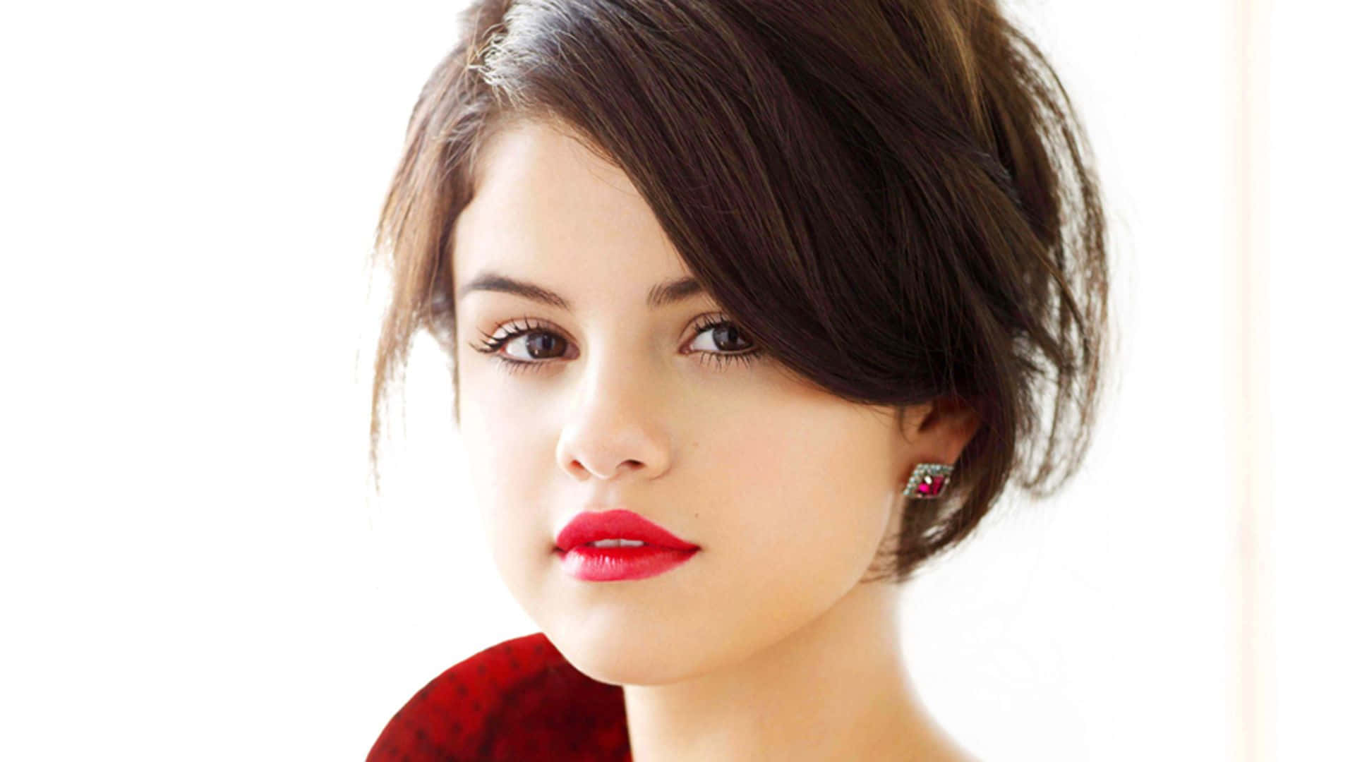 Selena Gomez 桌布 壁紙 — 一個特寫肖像，一位短黑髮、大膽紅唇、戴著閃亮耳環的女性，背景是明亮的白色。