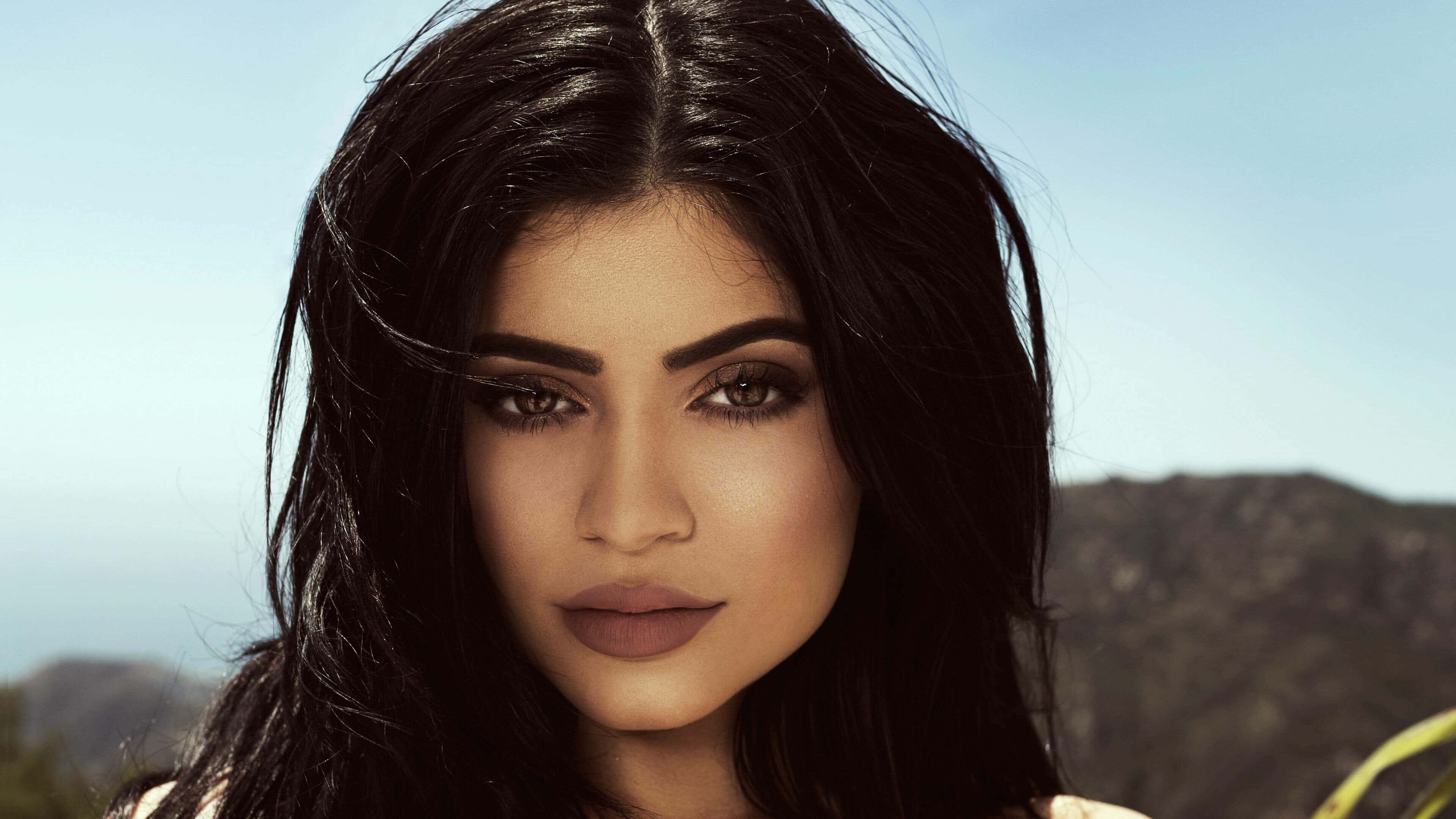 Kylie Jenner 4K 桌布 壁紙 — 一個特寫肖像，一位黑髮、妝容分明的女性，背景是明亮的日光下朦朧的山巒。