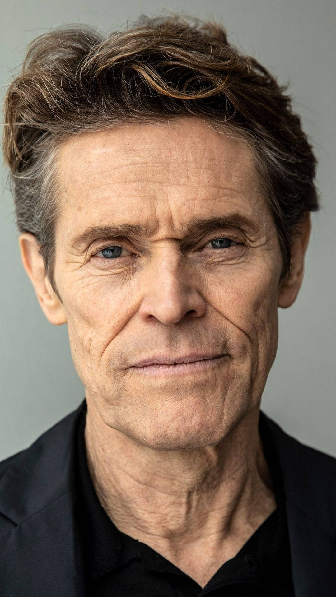 Willem Dafoe 桌布 壁紙 — 一個特寫肖像，一個有紋理棕色頭髮和強烈藍眼睛的男人，穿著黑色襯衫，背景為中性灰色。