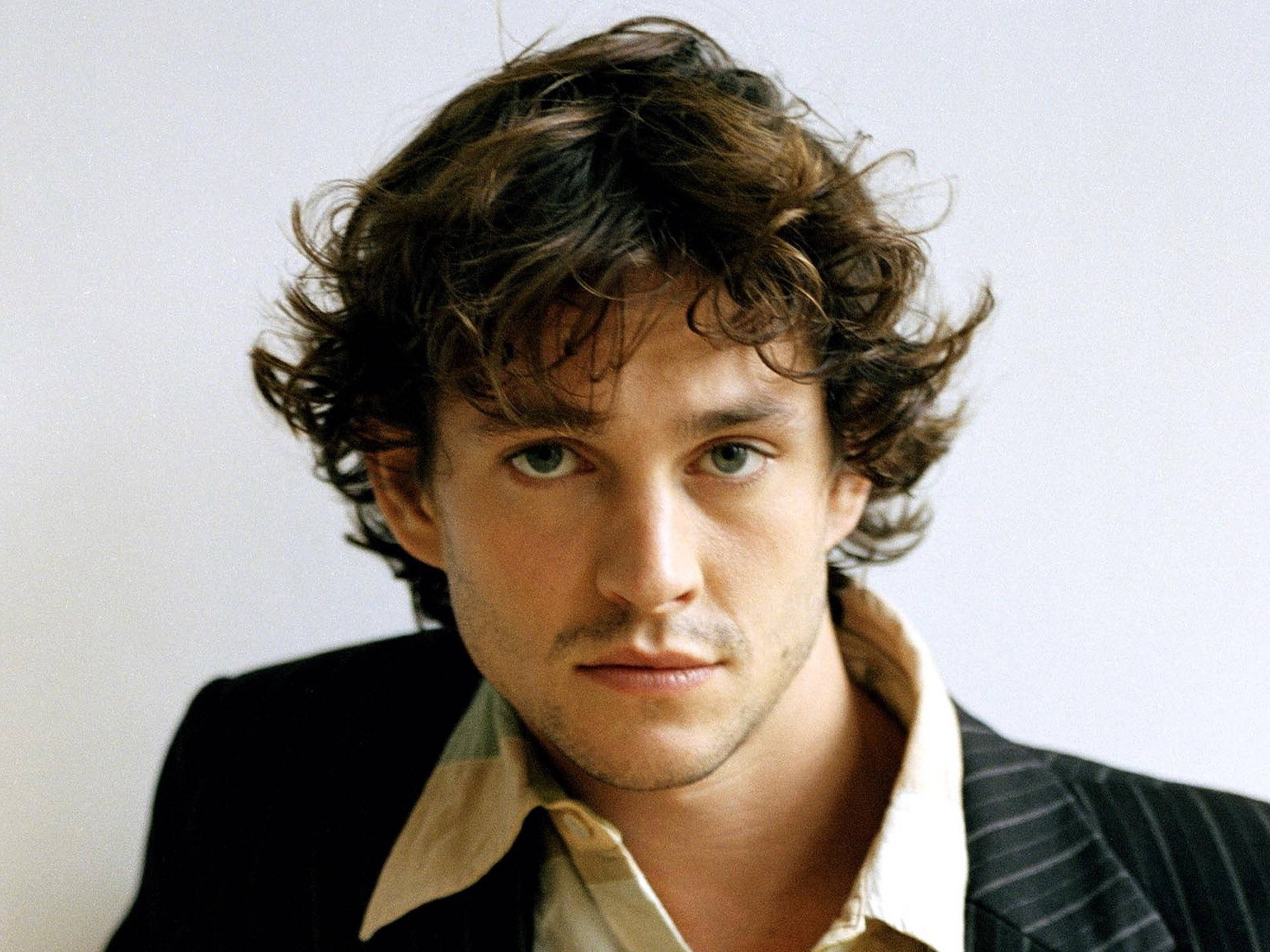 Hugh Dancy 桌布 壁紙 — 一個特寫肖像，一個男人有著凌亂的棕色頭髮和強烈的綠色眼睛，穿著細條紋西裝，背景是純白色。