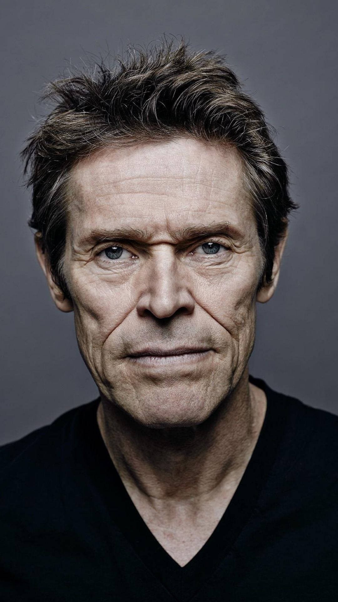 Willem Dafoe 桌布 壁紙 — 一個特寫肖像，一個飽經風霜的男人，擁有強烈的藍眼睛和凌亂的頭髮，背景為灰色。