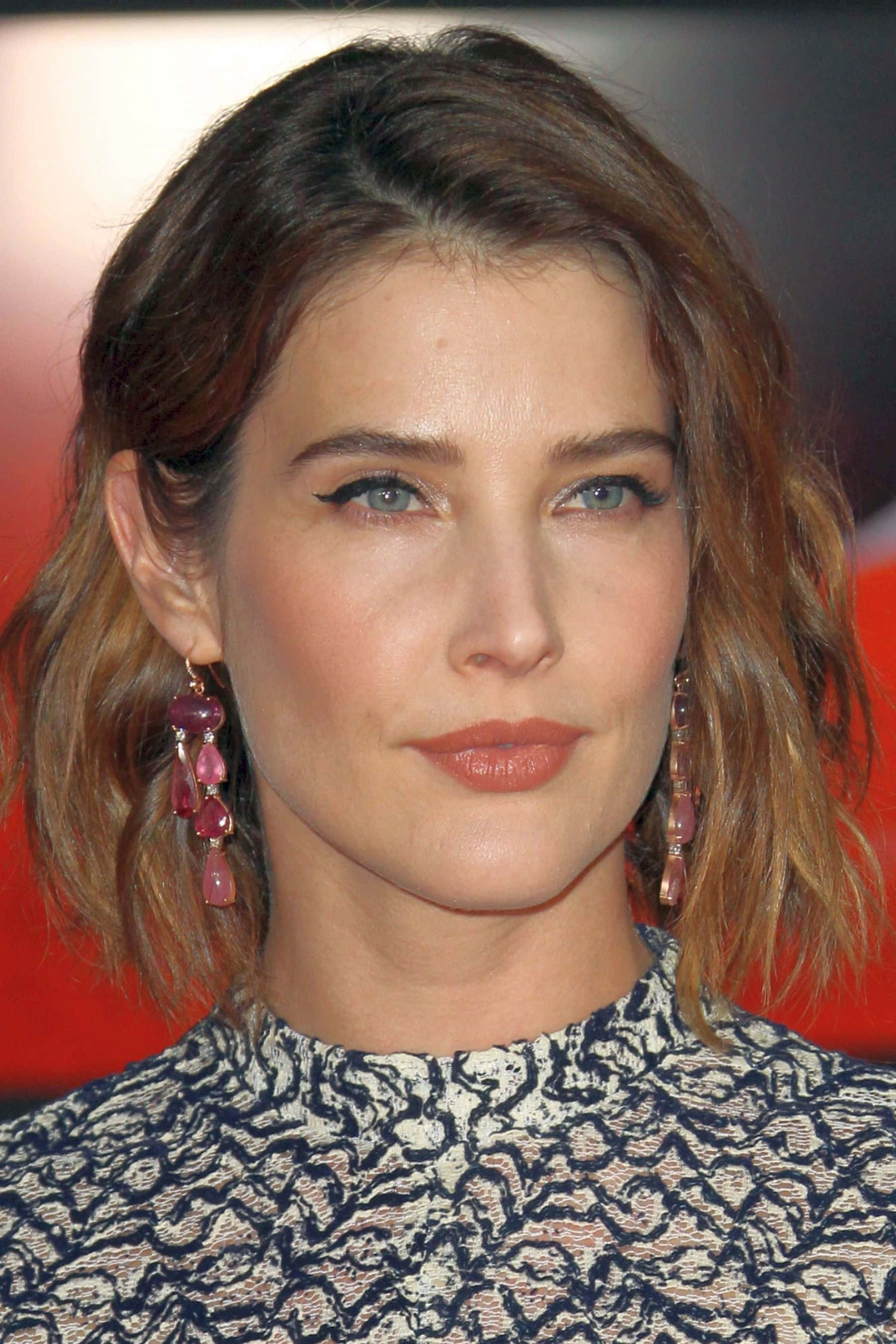 Cobie Smulders 桌布 壁紙 — 一個特寫肖像，一名擁有波浪棕色頭髮和引人注目的藍色眼睛的女性，戴著粉紅色吊墜耳環和圖案上衣，背景是模糊的紅色。