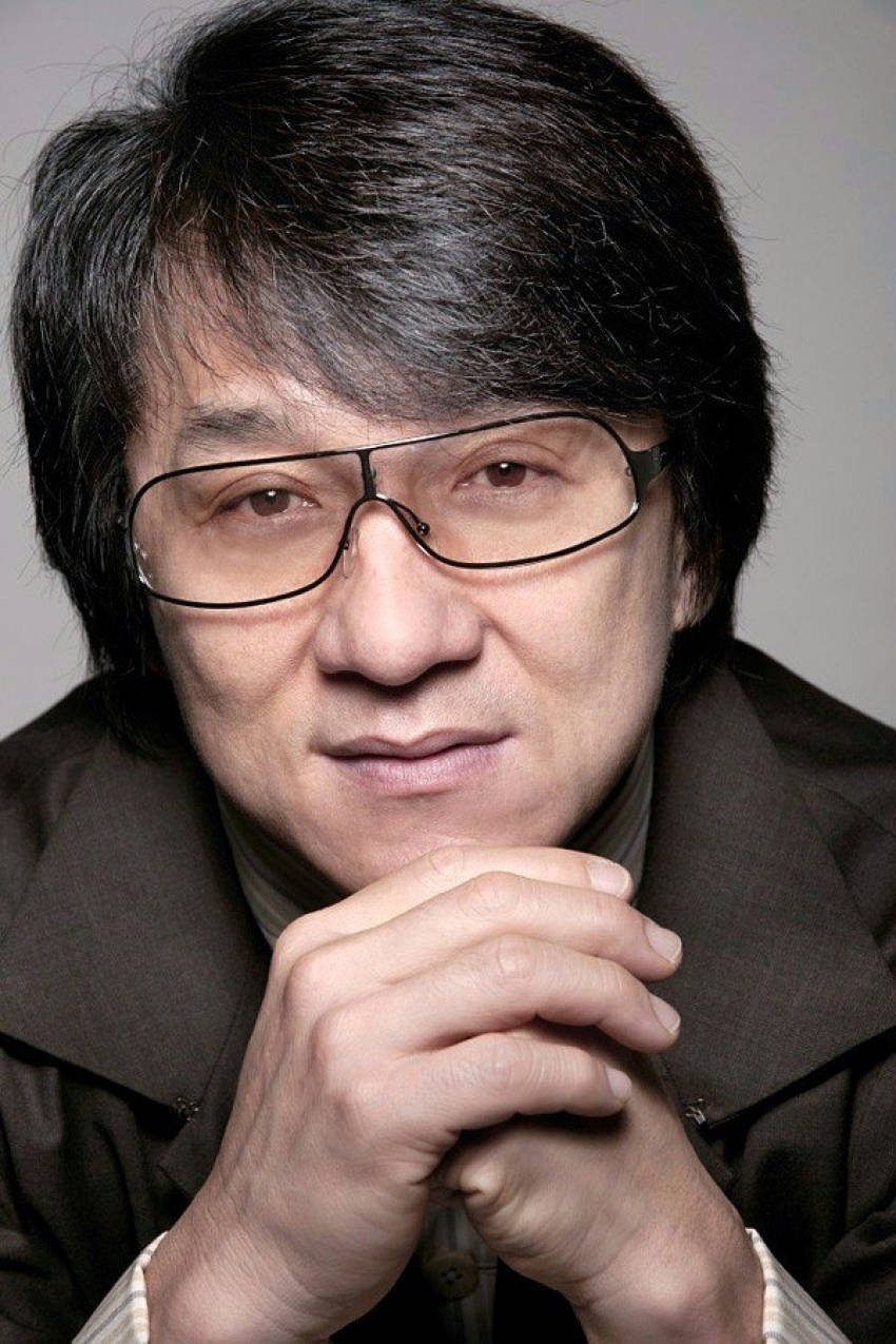 jackie chan 桌布 壁紙 — 一個特寫肖像，一名深色頭髮、戴眼鏡的男子，在灰色中性背景前，將交疊的雙手放在下巴下方。