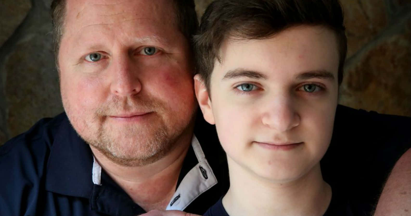 Father And Son 桌布 壁紙 — 一個特寫肖像，一名男子和一名年輕男子，藍色眼睛，對著鏡頭溫柔微笑，背景是石牆。
