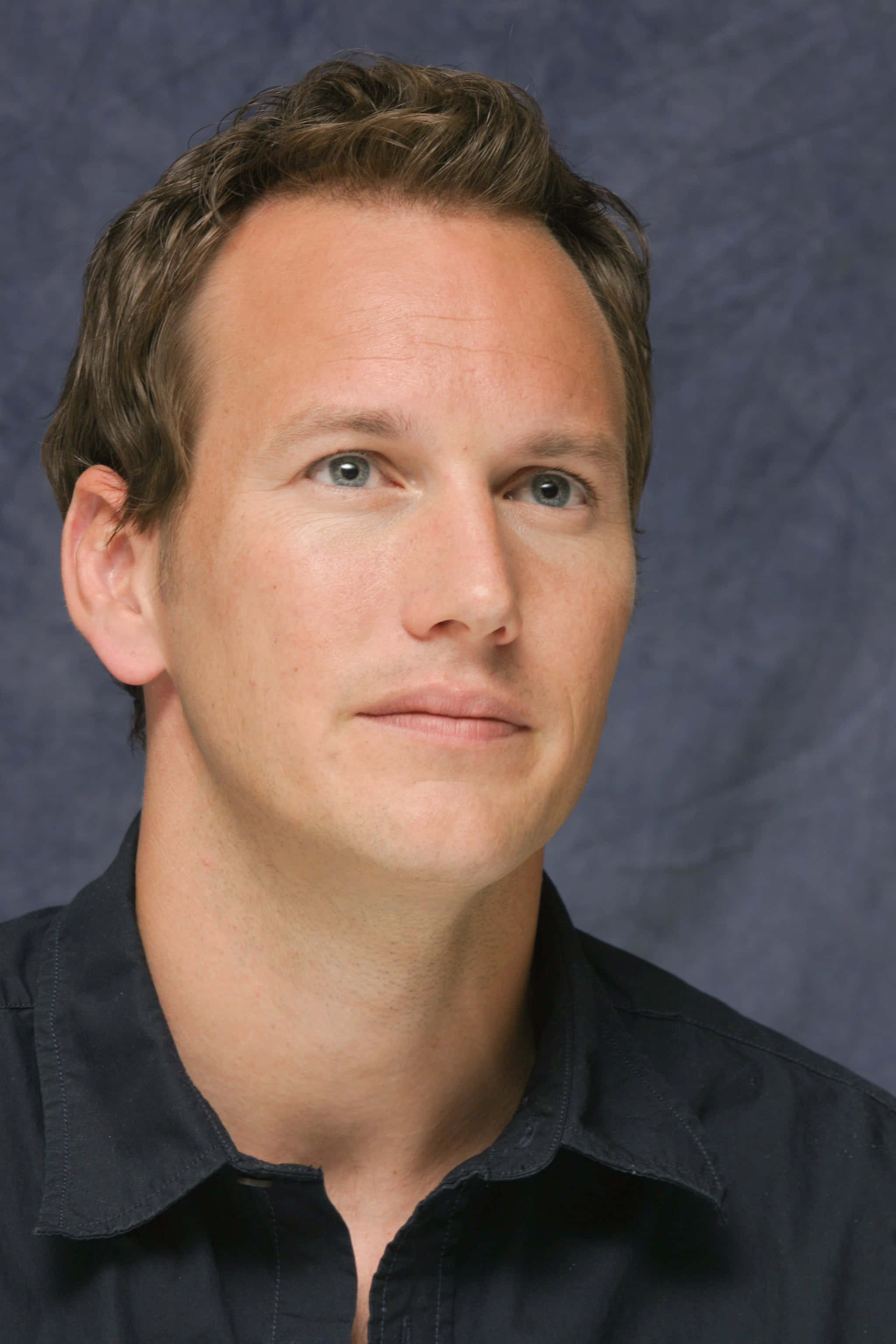 Patrick Wilson 桌布 壁紙 — 一個特寫肖像，一名男子擁有淺藍色眼睛和棕色頭髮，穿著深色襯衫，背景是帶紋理的藍色。