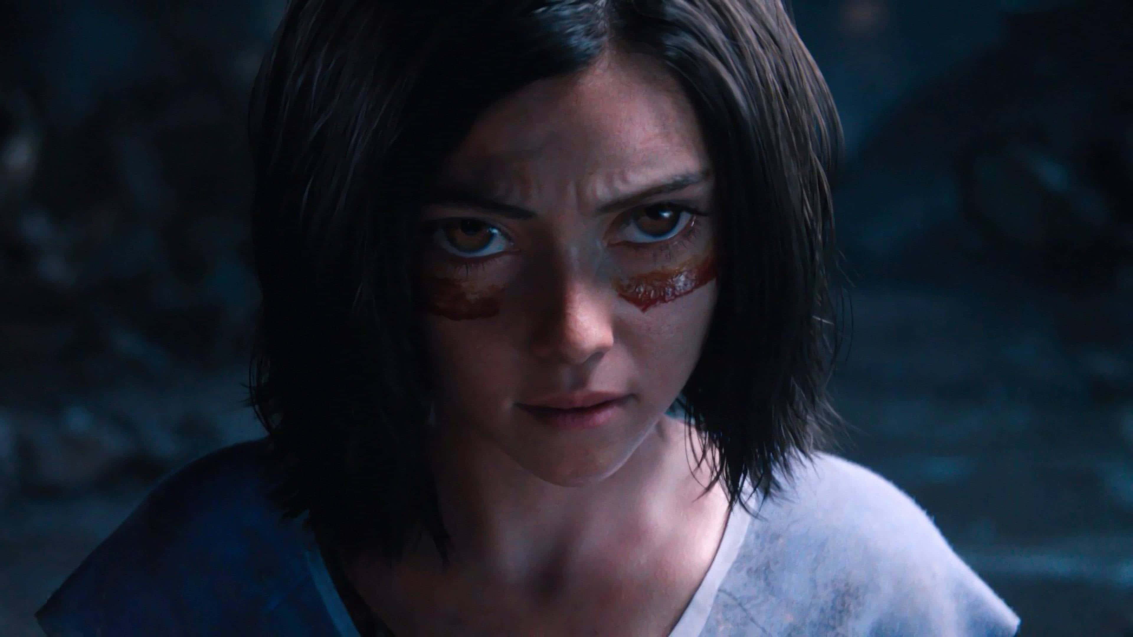 Alita Battle Angel 桌布 壁紙 — 一個特寫鏡頭，一位堅定的年輕女子，深色頭髮，臉頰帶血，在昏暗的洞穴中凝視。