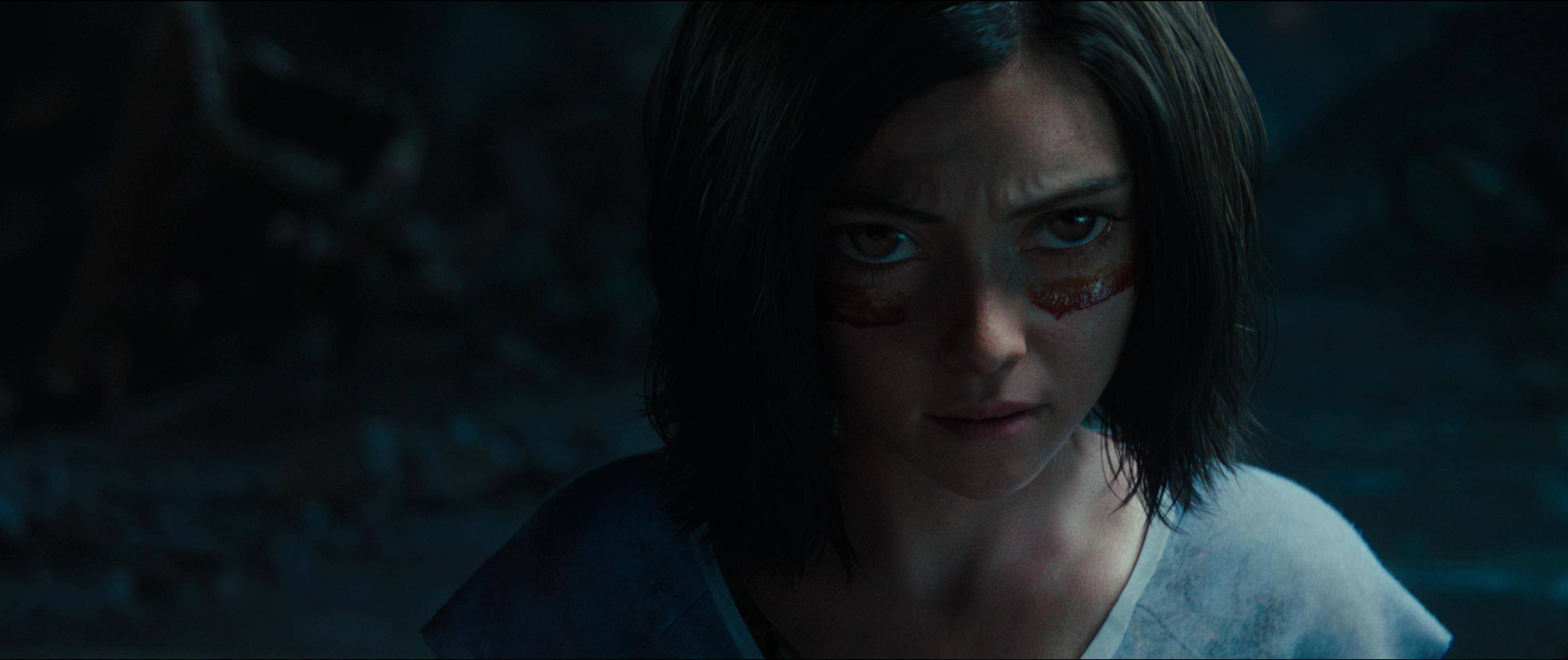 Alita Battle Angel 桌布 壁紙 — 一個特寫鏡頭，一位堅定的年輕女子臉頰帶血，在昏暗陰森的地下環境中凝視著。