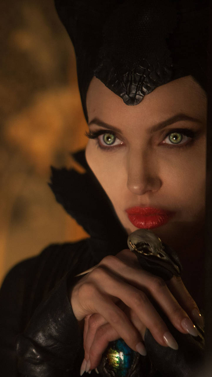 Maleficent 桌布 壁紙 — 一個特寫鏡頭，一位擁有引人注目的綠色眼睛和紅色嘴唇的女性，在溫暖模糊的背景前手持一根頂端有骷髏頭的權杖。