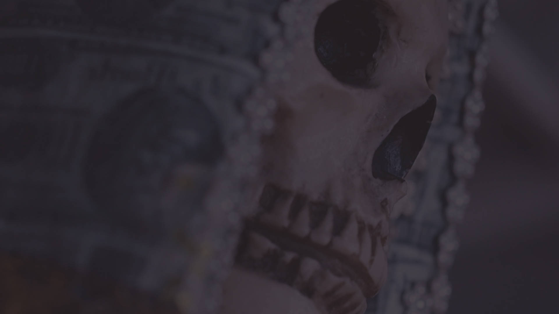 Santa Muerte 桌布 壁紙 — 一個特寫鏡頭，一個裝飾著珍珠和珠子的頭骨，在昏暗的燈光下放在一疊美元鈔票上。