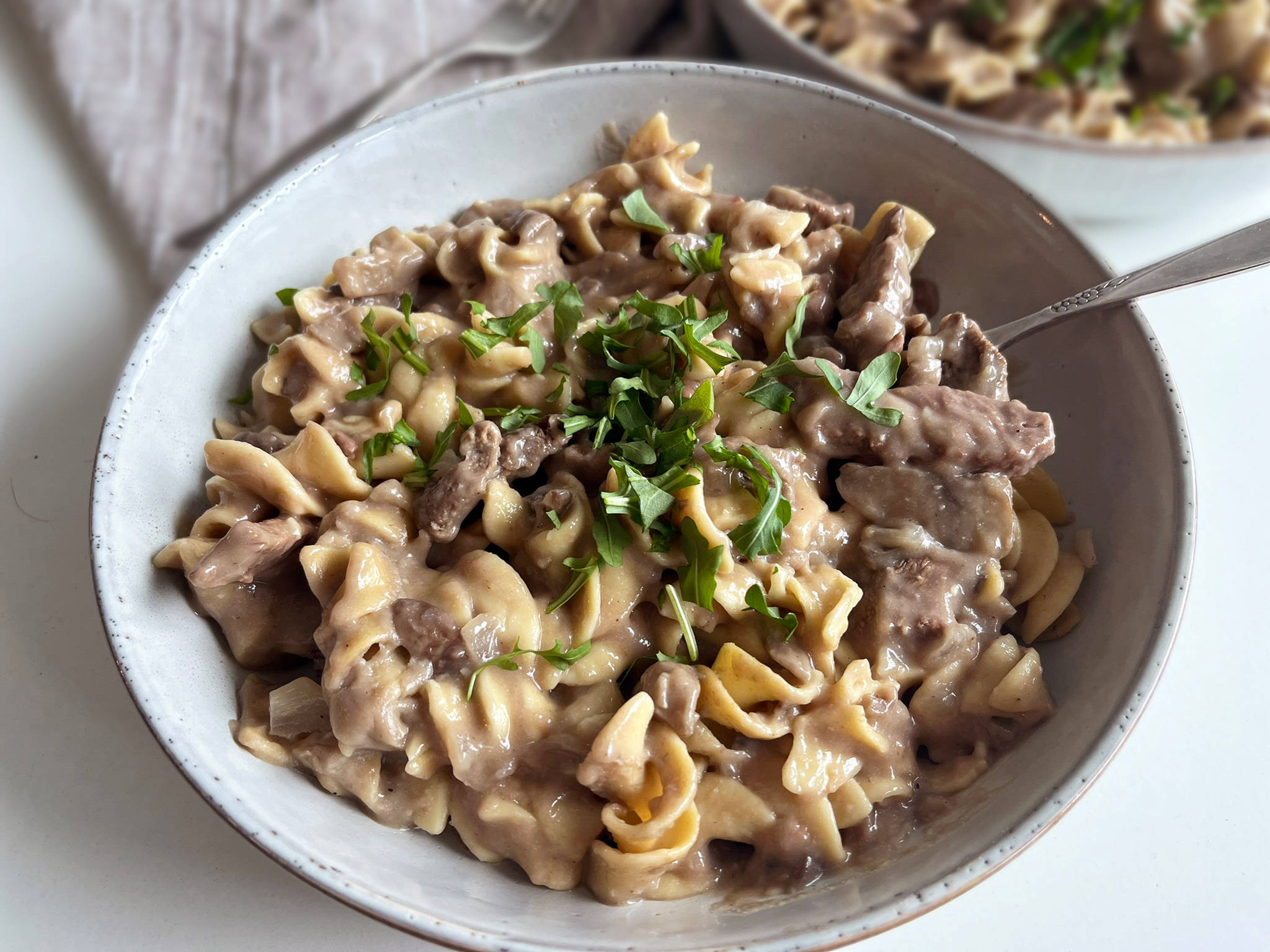 Beef Stroganoff 桌布 壁紙 — 一個特寫鏡頭，奶油牛肉斯特羅加諾夫配雞蛋麵和新鮮歐芹，放在一個有斑點的陶瓷碗裡。