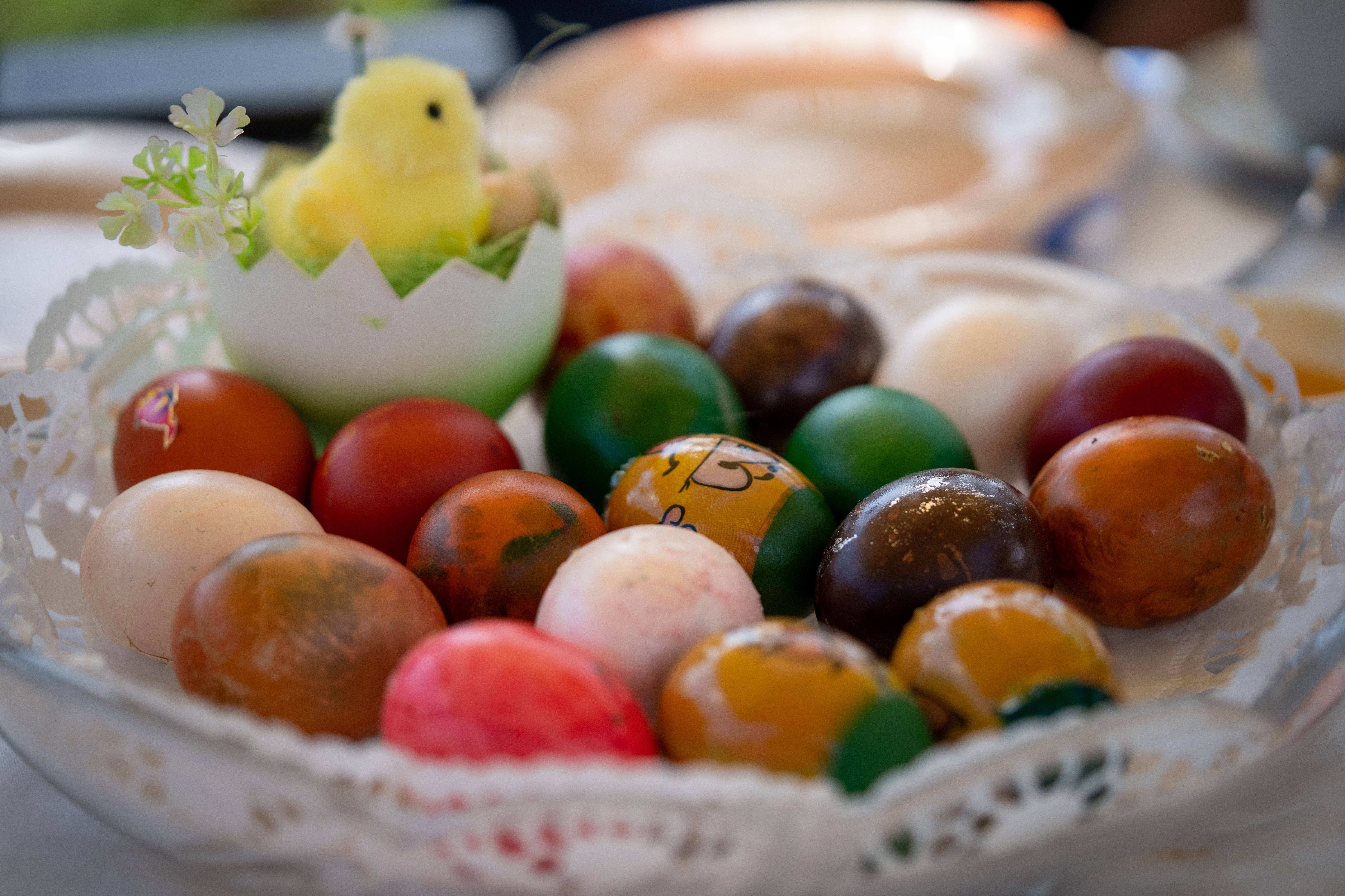 Happy Easter 桌布 壁紙 — 一個特寫鏡頭，展示一個鋪有蕾絲邊的碗中色彩繽紛的手繪復活節蛋，一隻黃色小雞從裂開的蛋中探出頭來。