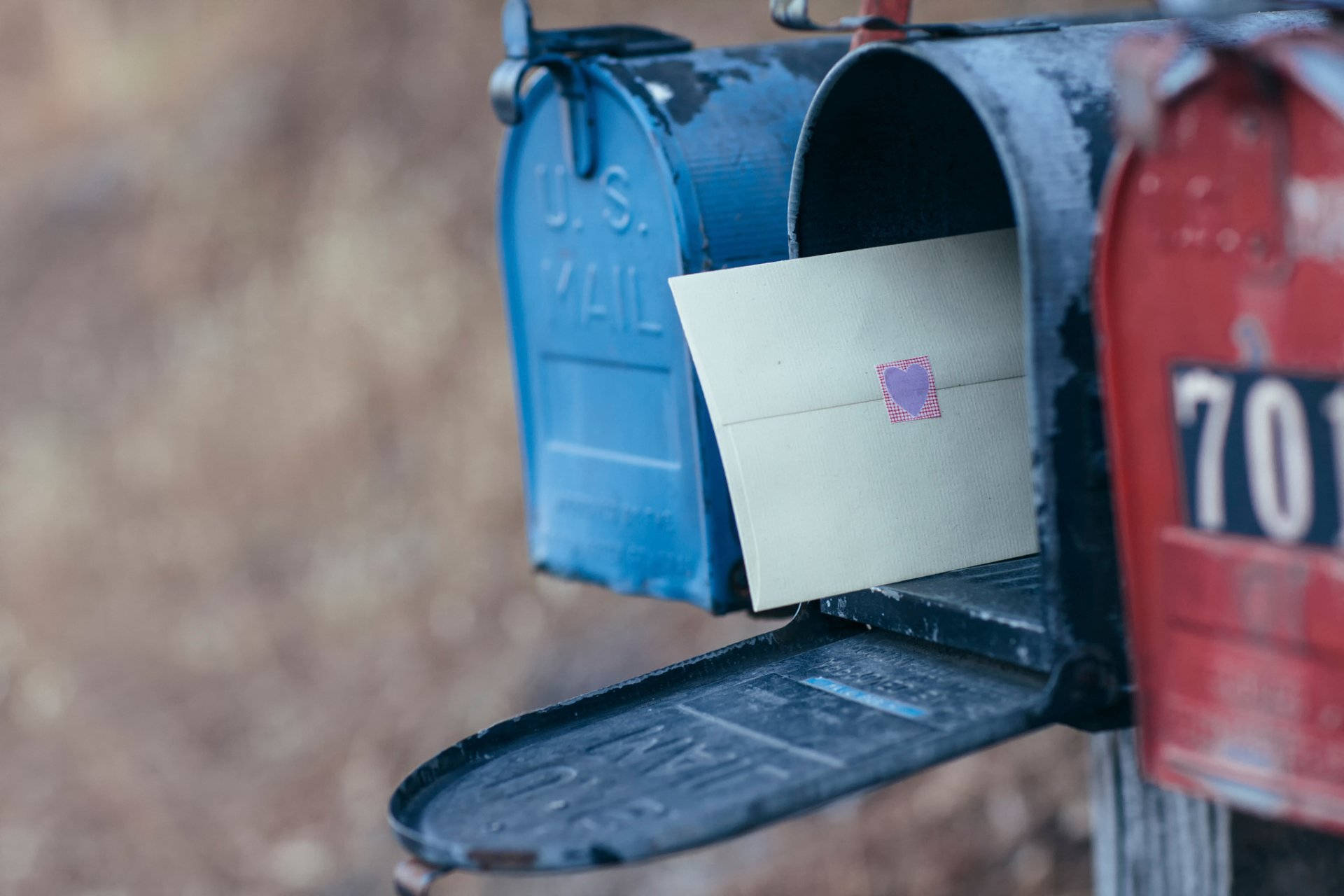 Post Office 桌布 壁紙 — 一個特寫鏡頭，展示一個飽經風霜的藍色郵筒，一個奶油色信封露出一枚紫色心形郵票。