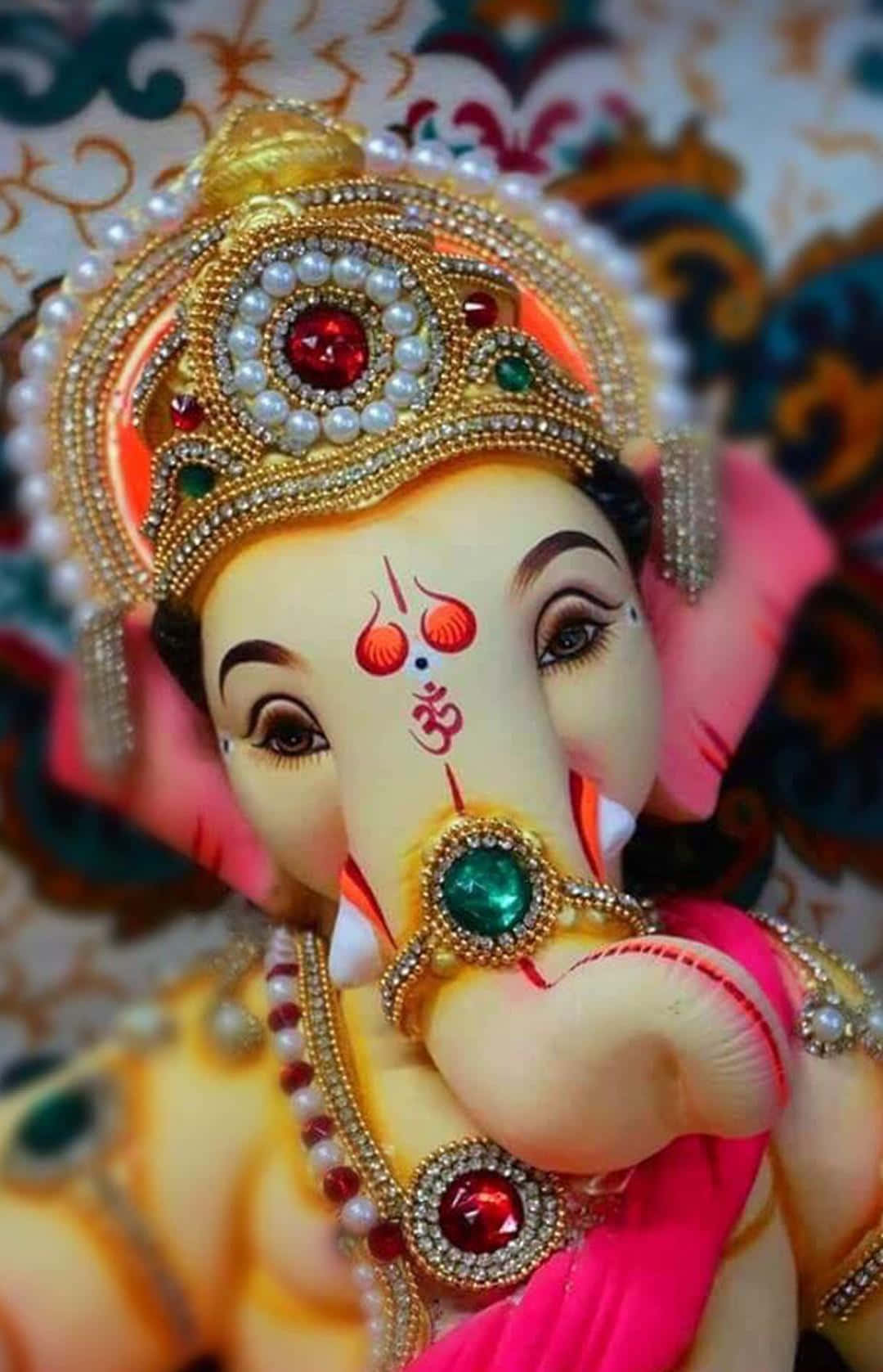 Ganesh 桌布 壁紙 — 一個特寫鏡頭，展示一尊裝飾華麗的象頭神像，戴著金色皇冠珠寶，身穿粉紅色服飾，背景帶有圖案。