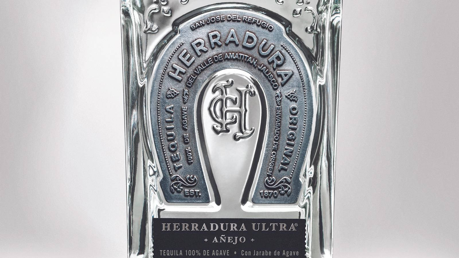 手機 桌布 壁紙 — 一個特寫鏡頭，展示一瓶清澈玻璃瓶裝的 Herradura Ultra Añejo 龍舌蘭酒，瓶身帶有精緻的銀色馬蹄鐵標誌，背景為柔和的灰色。