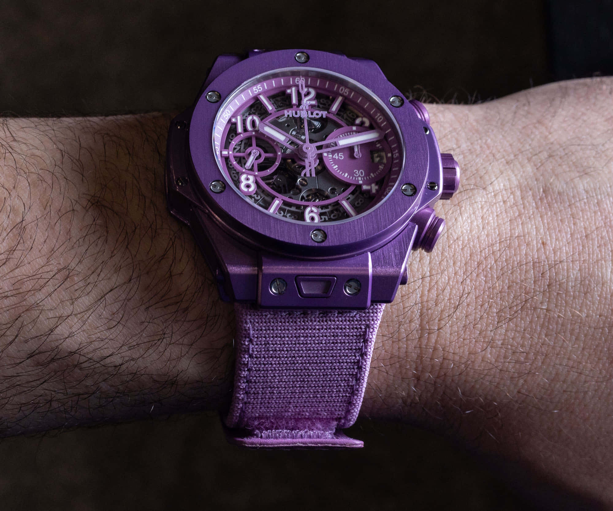 Purple Accessories 桌布 壁紙 — 一個特寫鏡頭，展示一隻鮮紫色Hublot手錶配以織物錶帶，戴在毛茸茸的手腕上，背景為深色。