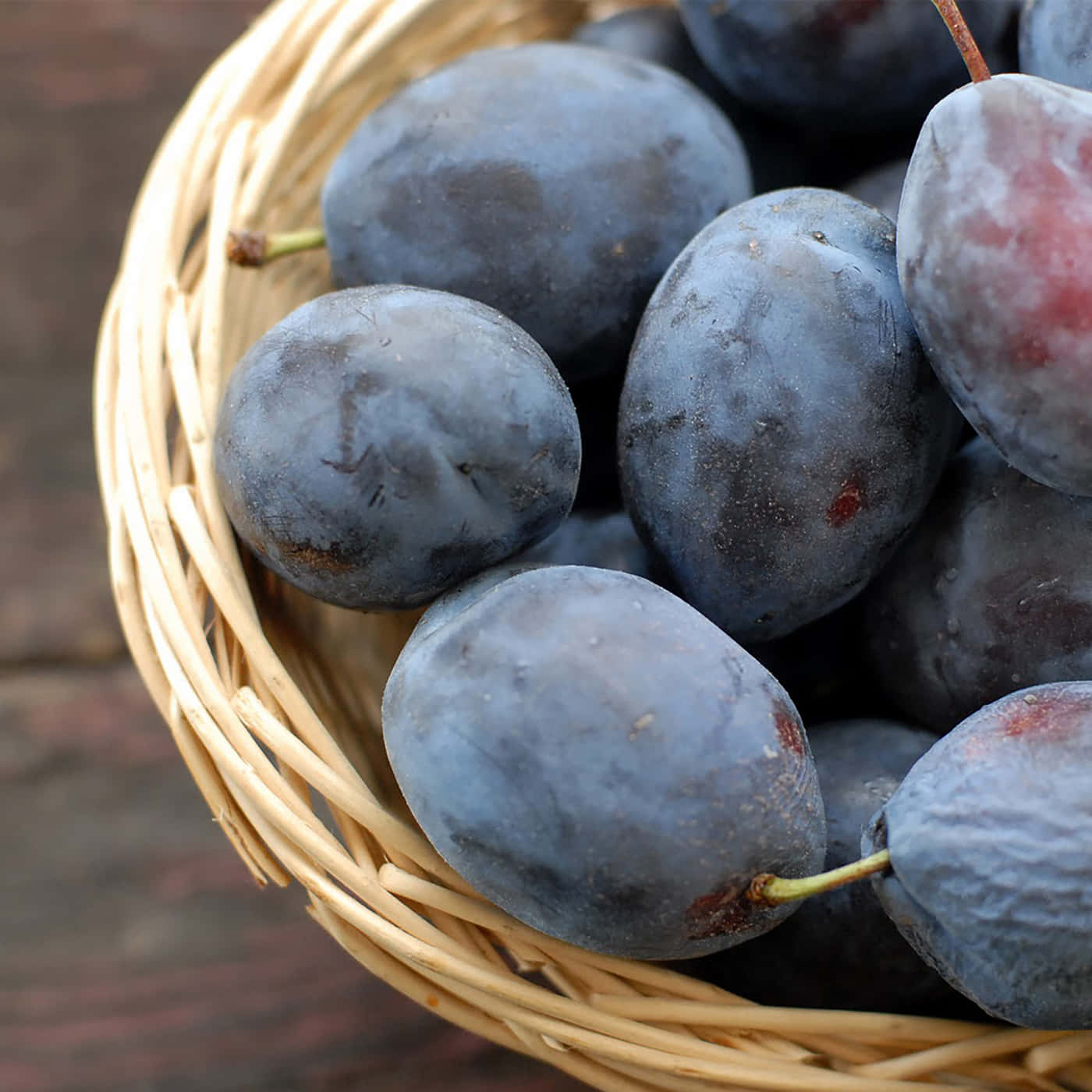 Fruit 桌布 壁紙 — 一個特寫鏡頭，展示了一個編織柳條籃子裡裝滿了飽滿的深紫色李子，放在質樸的木質表面上。