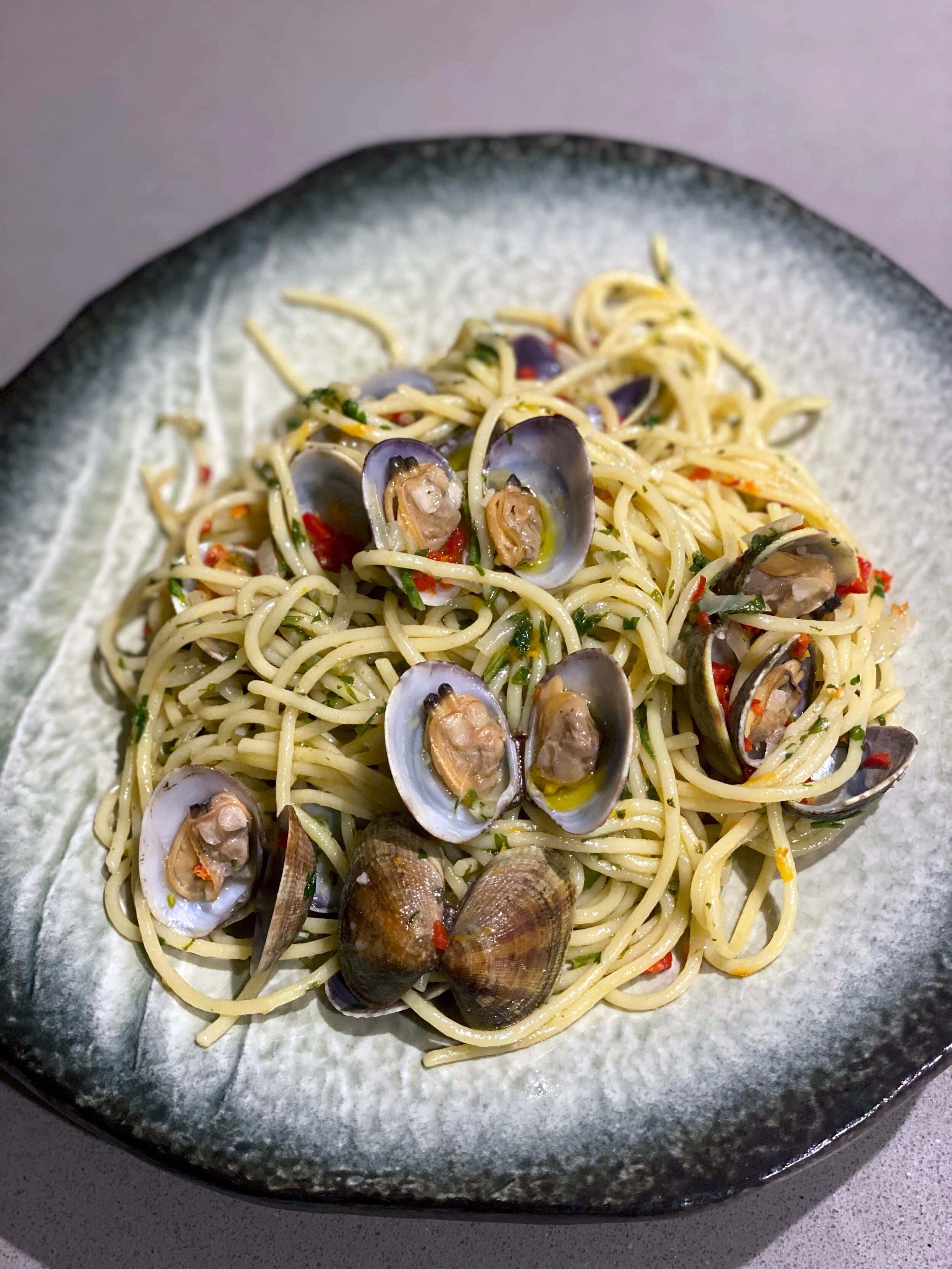 Clam 桌布 壁紙 — 一個特寫鏡頭，展示放在有紋理的灰色碗中的蜆肉義大利麵，上面裝飾著香草和紅椒。