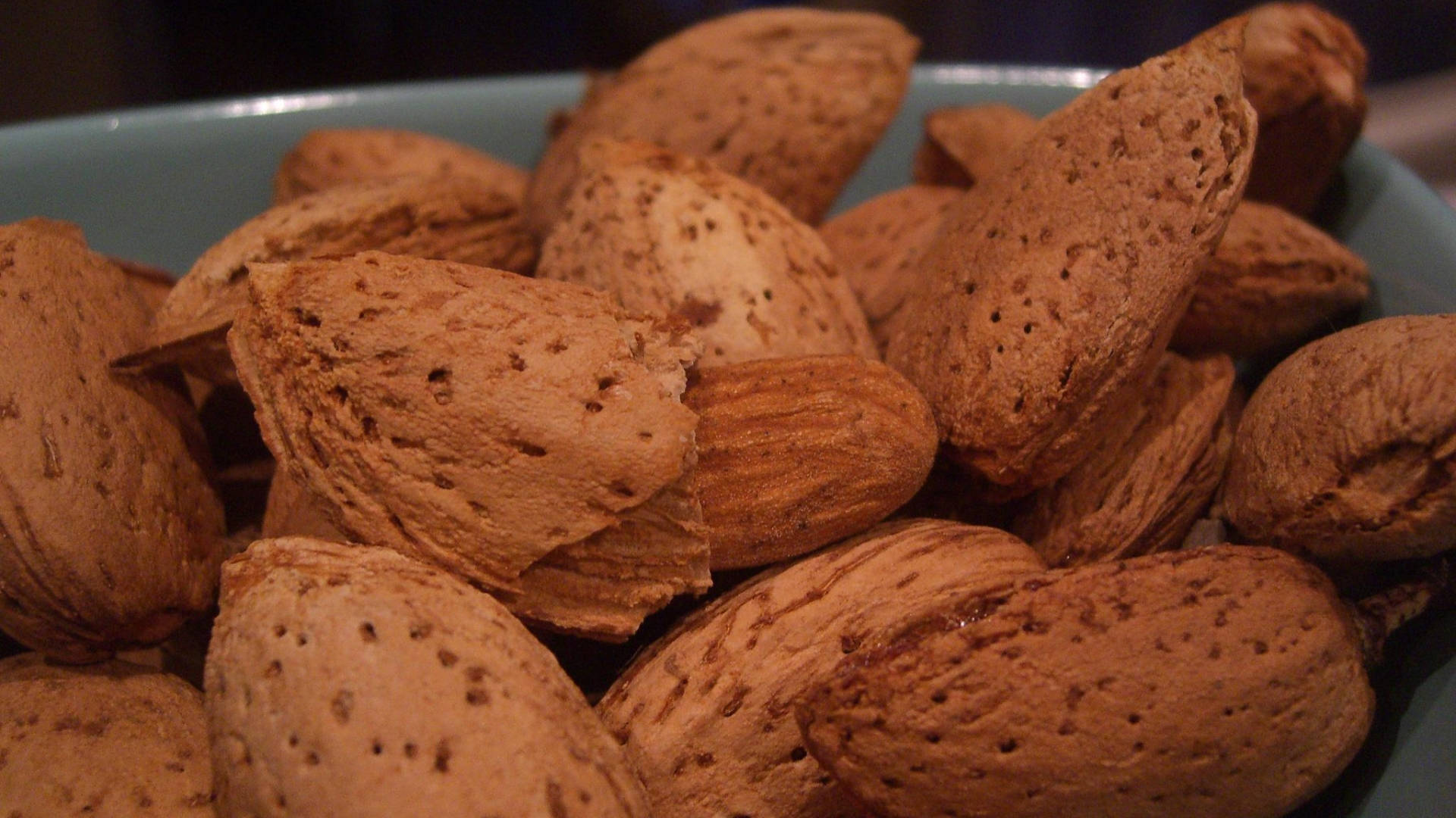 Almond 桌布 壁紙 — 一個特寫鏡頭，展示放在青綠色碗中的烤杏仁，溫暖的燈光突顯其紋理外皮。