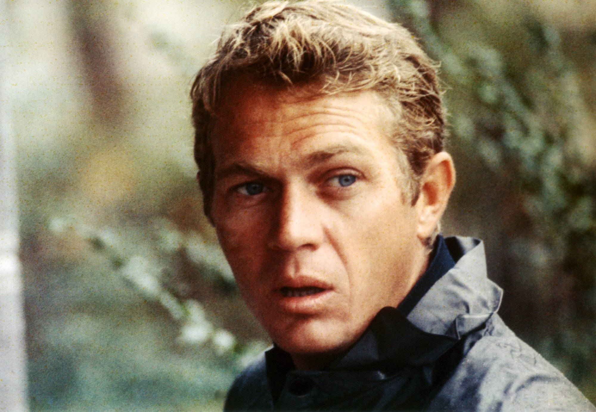 actor 桌布 壁紙 — 一個特寫鏡頭，捕捉一名擁有深邃藍眼睛和凌亂頭髮的男子，在模糊的綠色樹葉背景下直視鏡頭。