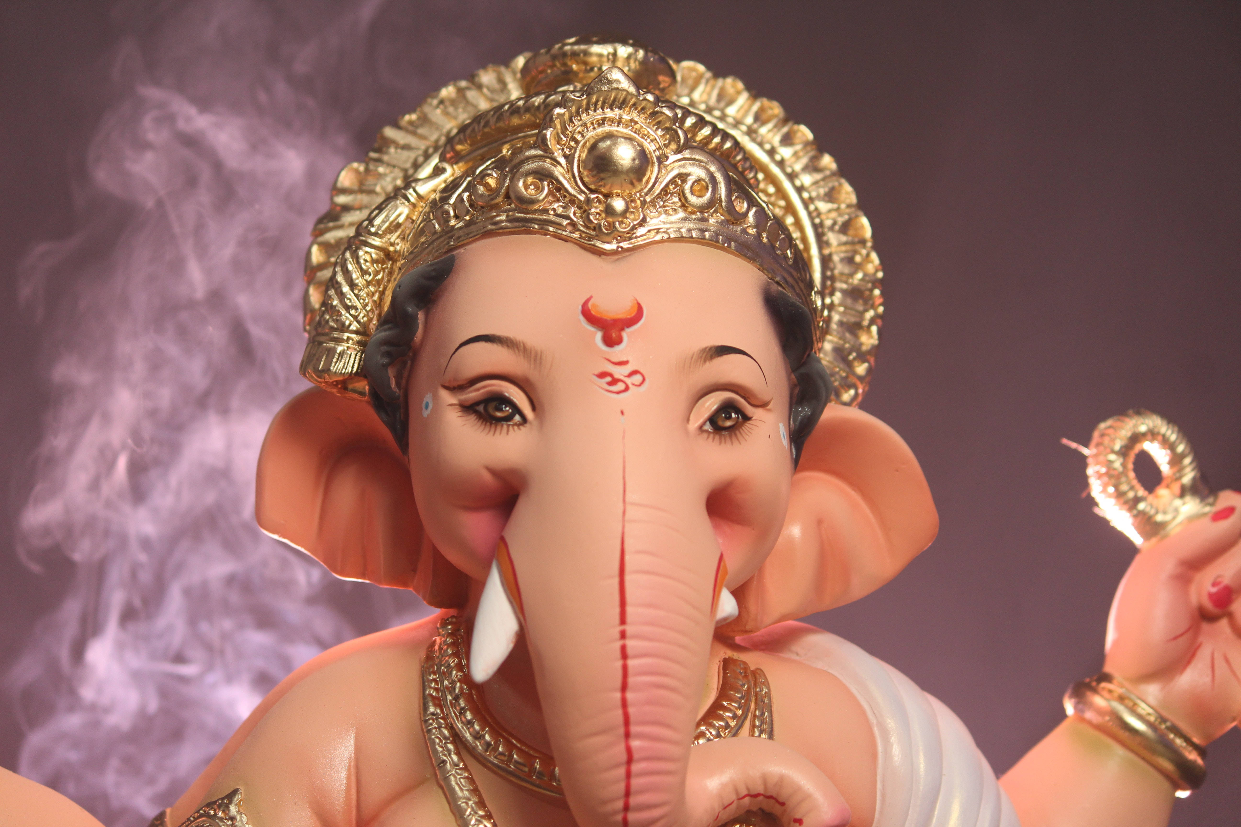 ganesh 4K 桌布 壁紙 — 一個特寫鏡頭，描繪一尊細節精緻的象神雕像，頭戴金色皇冠，帶有紅色標記，手持發光環，背景為煙霧瀰漫的紫色。