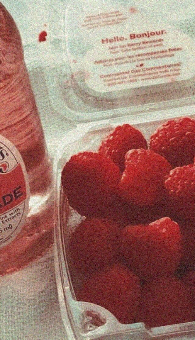 Soft Red Aesthetic 桌布 壁紙 — 一個特寫鏡頭，新鮮紅覆盆子放在透明塑膠盒內，旁邊有一支帶標籤的粉紅色飲料瓶。