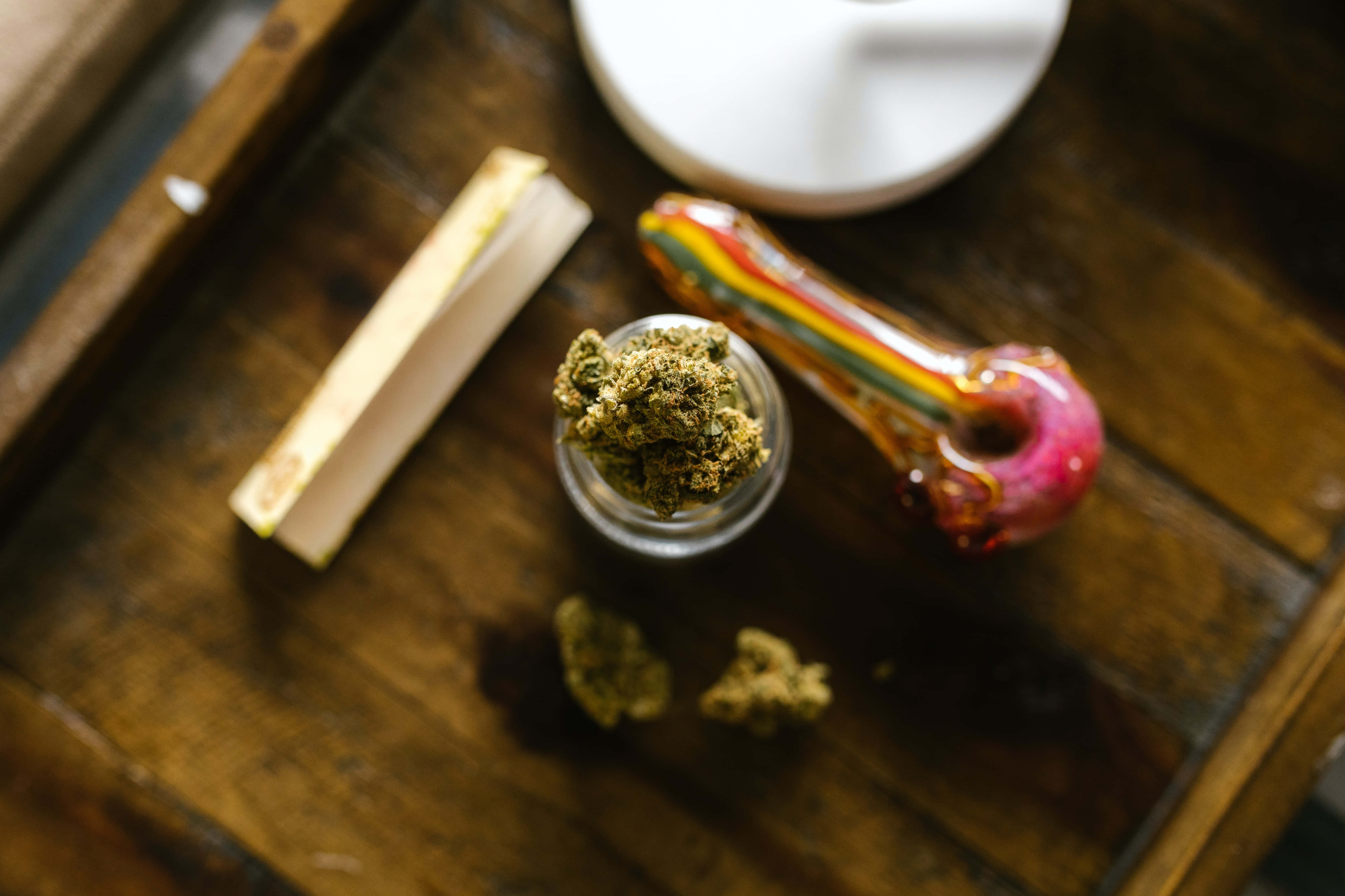 Weed Blunt 桌布 壁紙 — 一個特寫鏡頭，顯示一個鄉村風格木托盤上，一個罐子裡的大麻花蕾旁邊放著彩虹玻璃煙斗和捲煙紙。